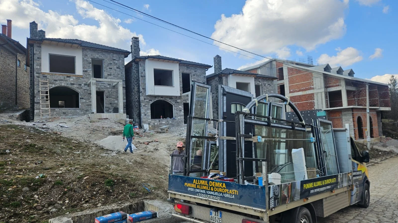 Construction site with three new buildings in progress on a sloped terrain under a partly cloudy sky. A truck with glass panels and construction materials is parked in the foreground. Two workers and one man are visible on site, with one worker weari