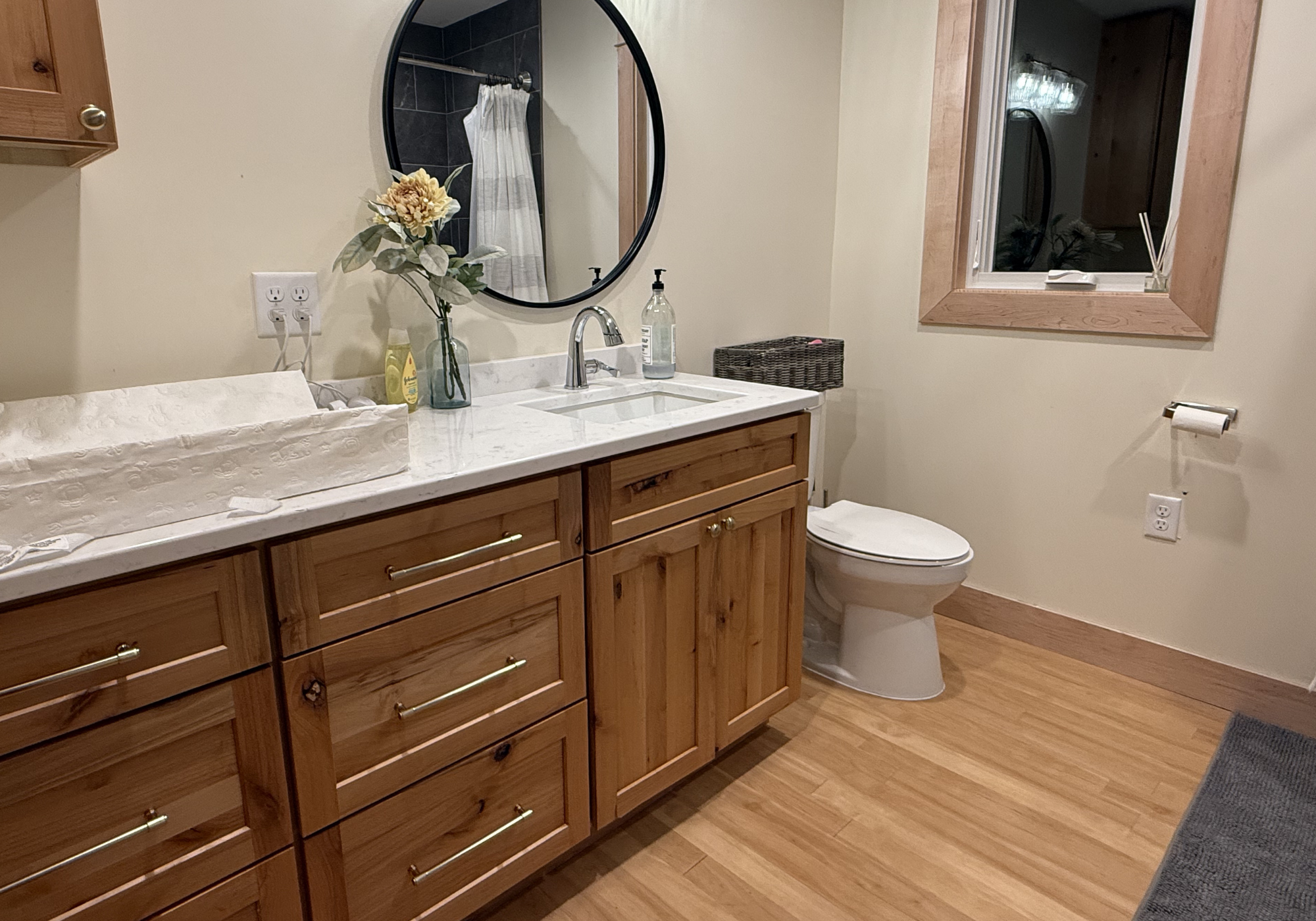 Bathroom with wooden vanity, quartz countertop, oval mirror, flower vase, soap dispenser, window, and toilet.
