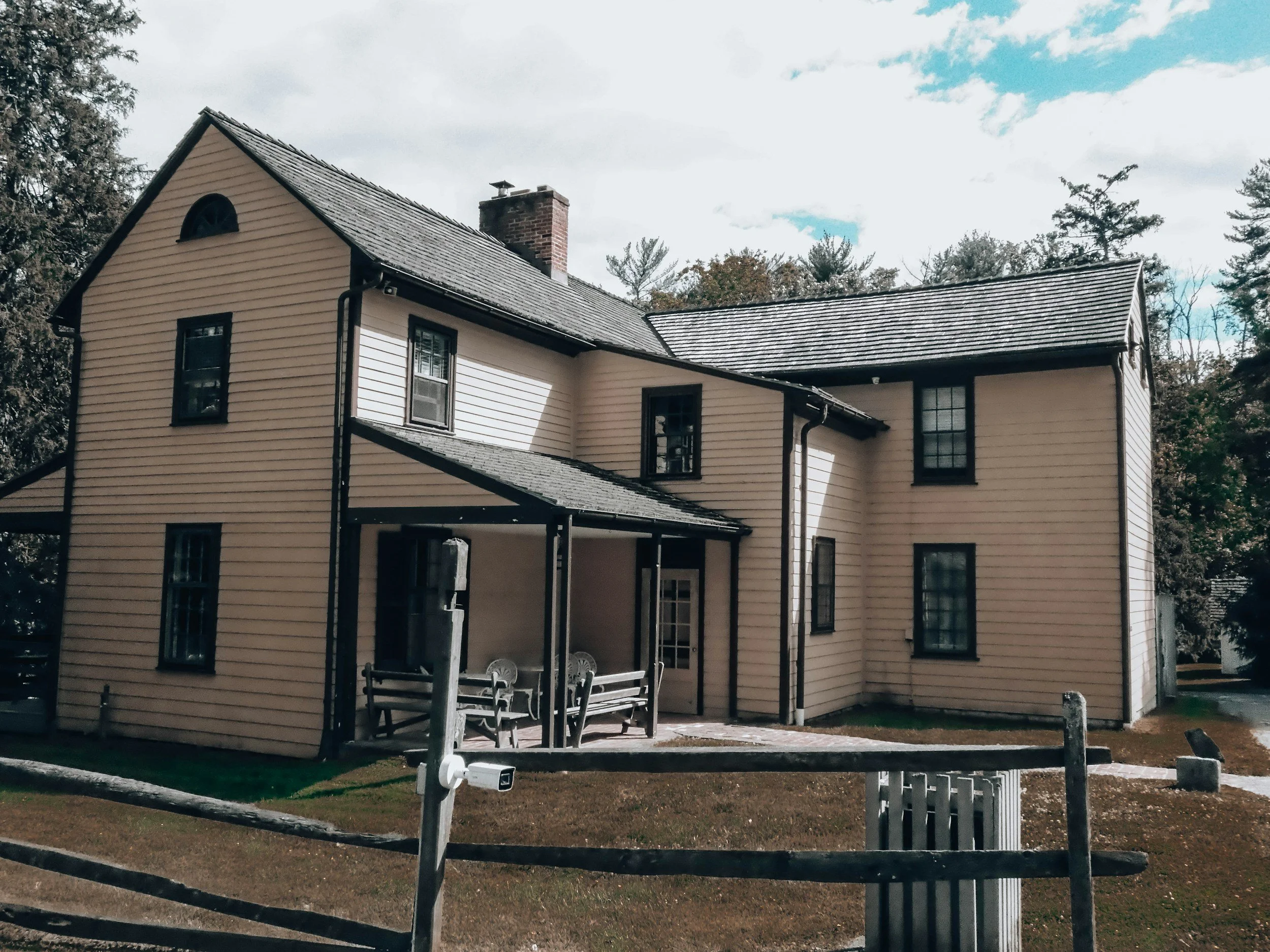A cream-colored, multi-story house with black window frames and a brick chimney. It has a sloped roof with asphalt shingles and a small arched window near the peak. There is an outdoor area with benches, a wooden railing, and some trees in the background, under a partly cloudy sky.