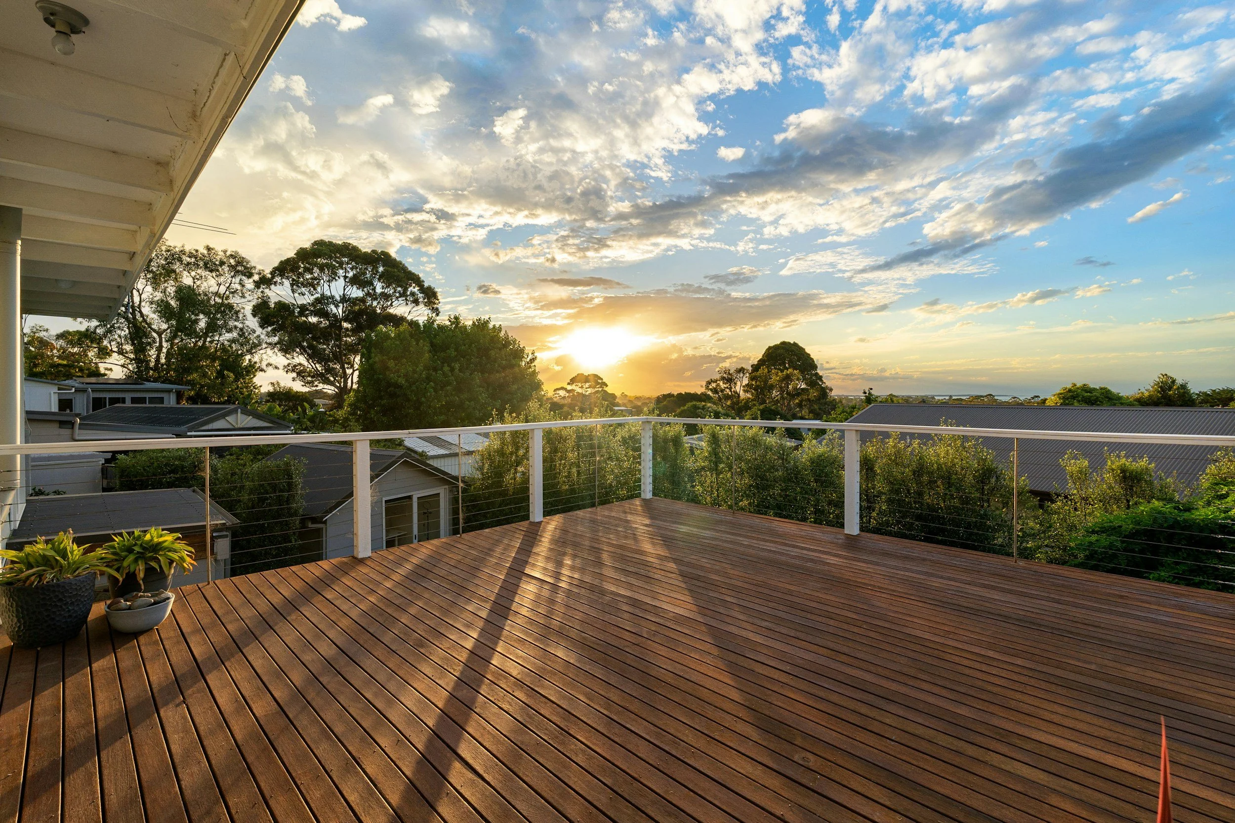 Sunset view from a wooden balcony with potted plants, overlooking houses, trees, and a partly cloudy sky.