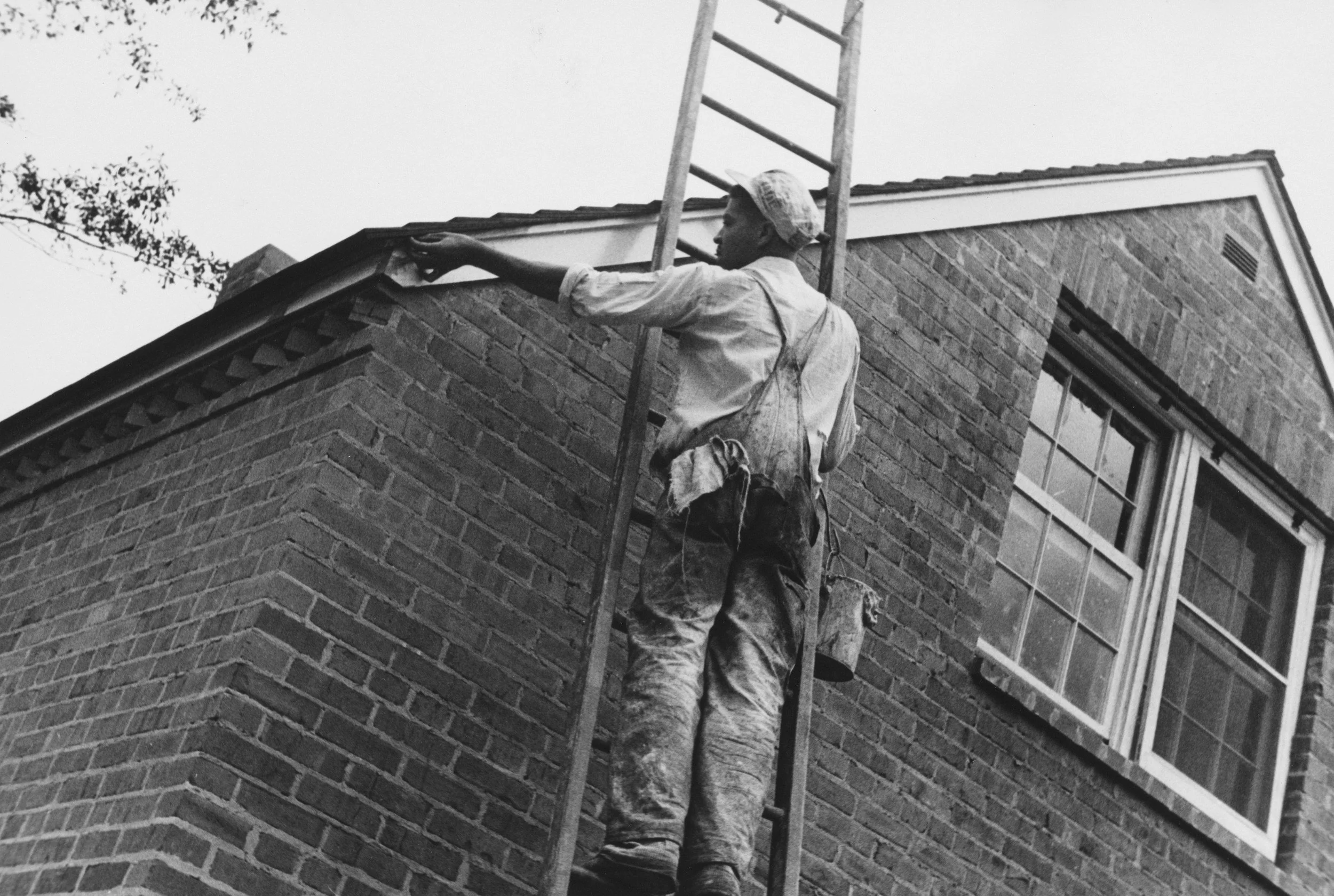 A person standing on a ladder painting the edge of a roof on a brick house.