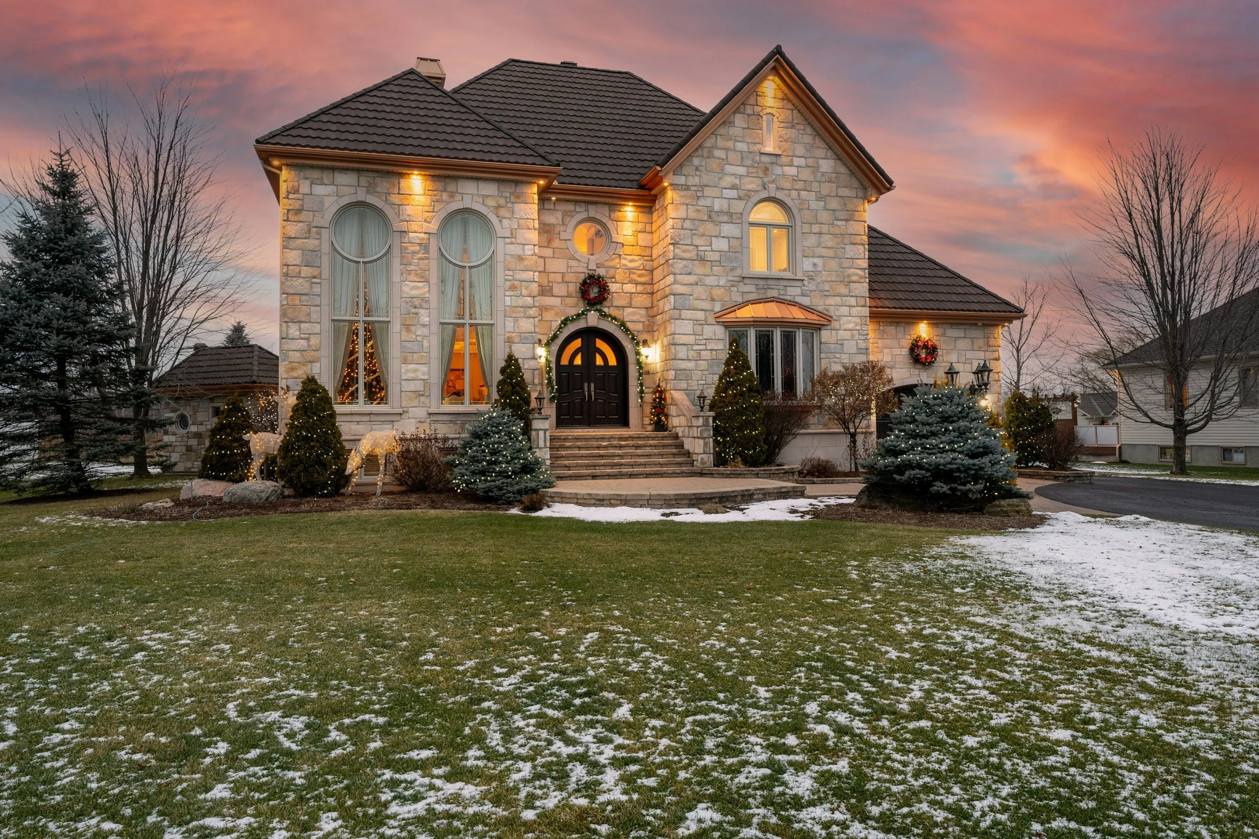 A large stone house decorated with Christmas wreaths and lights during dusk, with a manicured lawn and patches of snow.