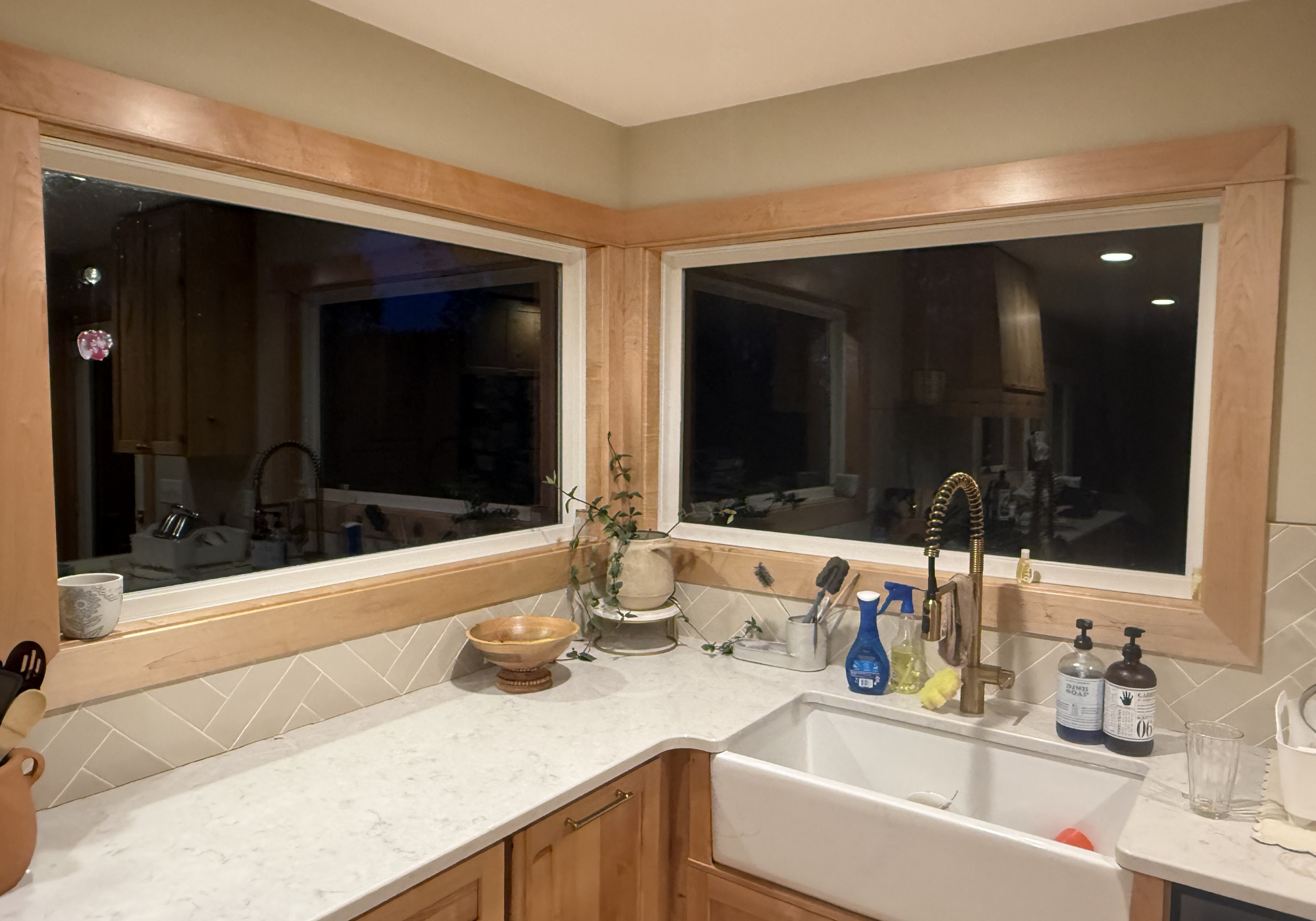 A kitchen corner with a white countertop, a sink, and big corner windows that offer a view outside. Soap bottles, a yellow sponge, and cleaning tools on the counter. Window trim is clear maple, and a small plant with green leaves on the counter.
