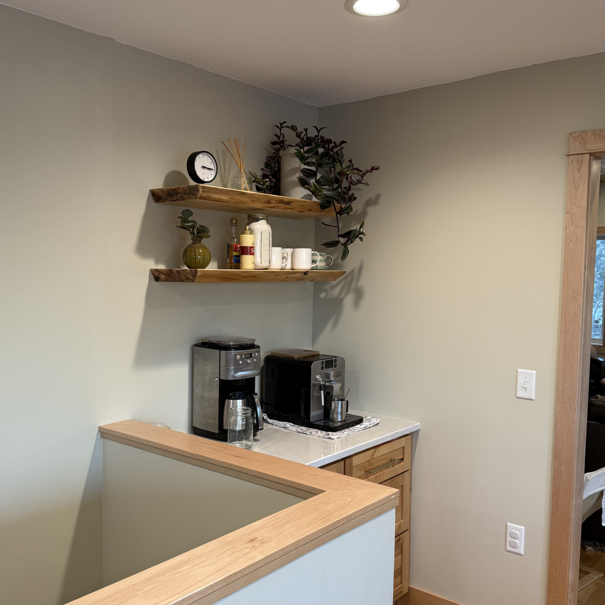 A  with a coffee bar and floating shelf on a gray wall decorated. Below the shelf, there are white cups, a glass, and a coffee maker on the counter. A wooden drawer cabinet is visible.