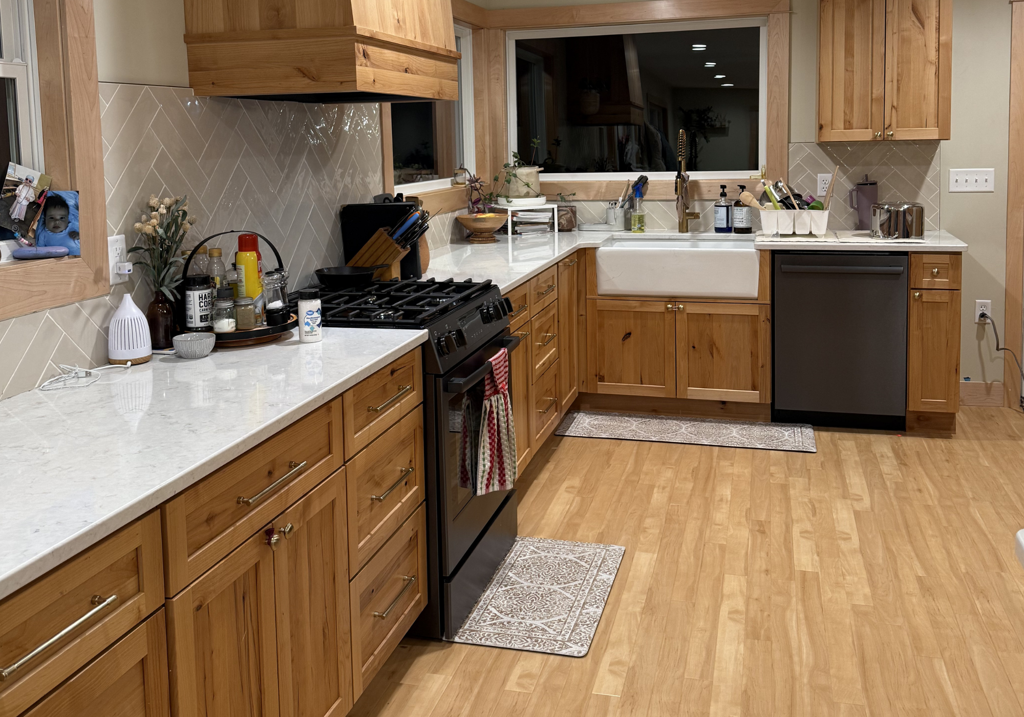Kitchen with wooden cabinets, white countertops, a white farmhouse sink, and a black dishwasher. Items on the counter include a small plant, containers, dishes, soap dispenser, and various kitchen tools. There is a window above the sink and a patterned rug on the wooden floor.