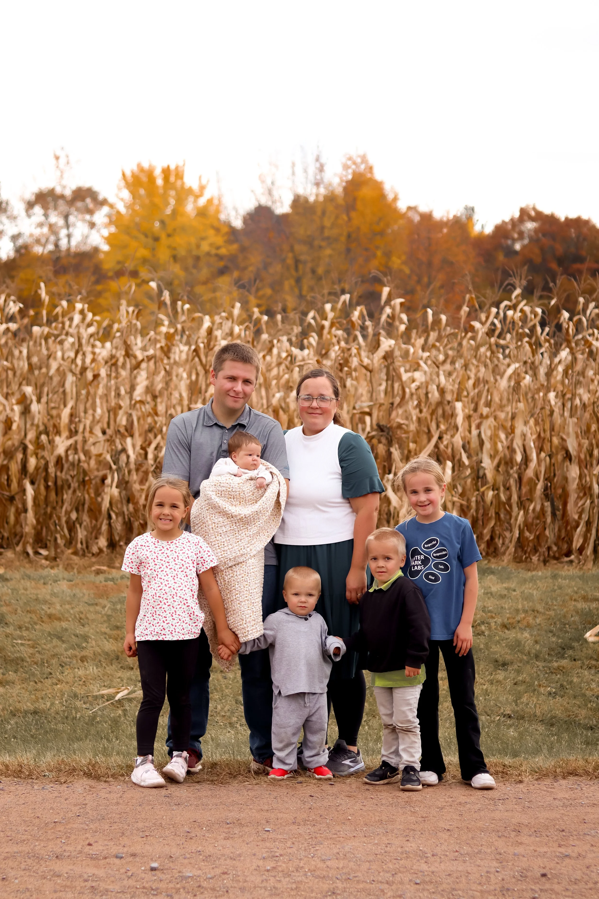 A group of children and two adults standing in front of a cornfield during autumn with trees in the background.