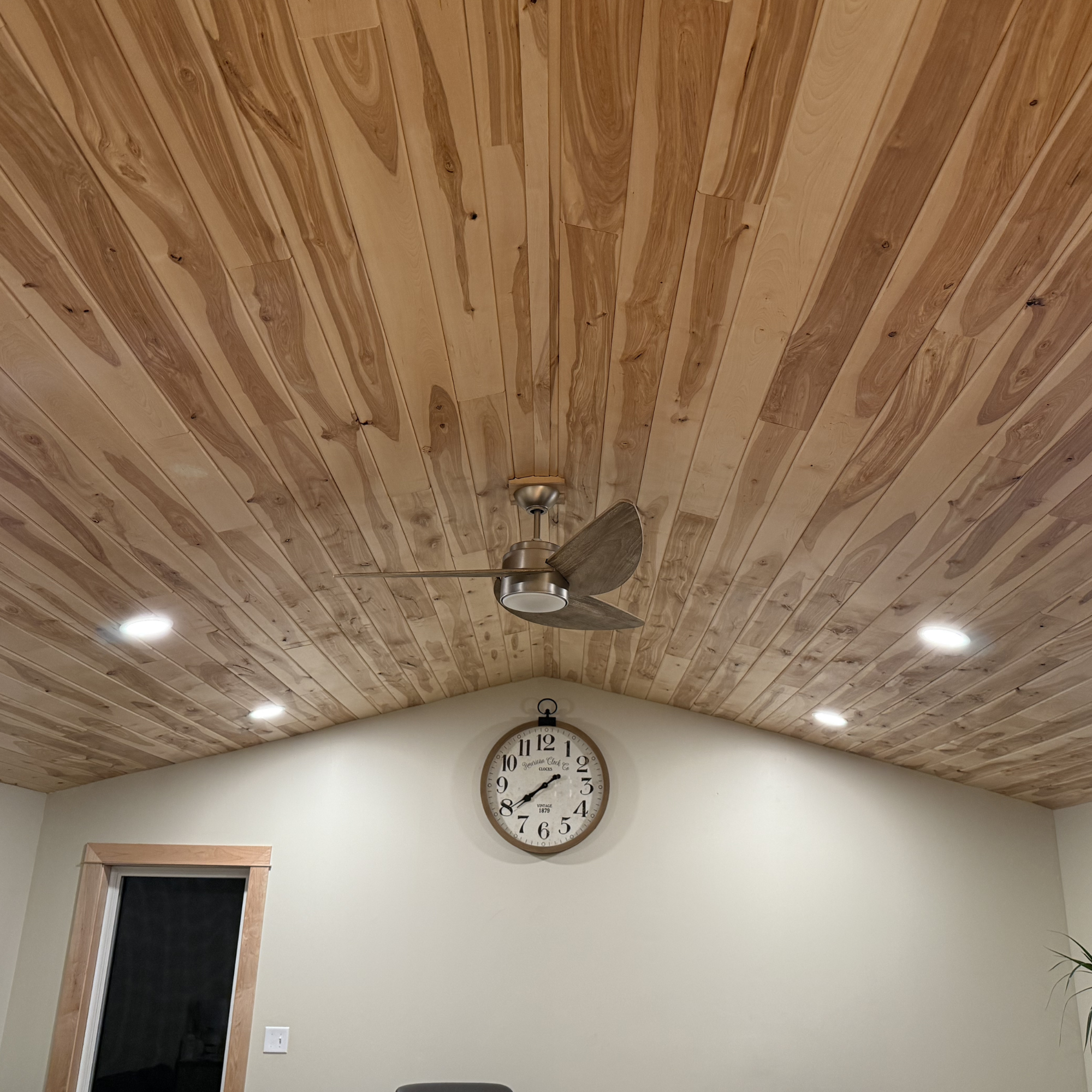 Interior view of a wood tongue and groove ceiling with a fan and a wall clock showing the time 8:29, along with a window and a light switch.