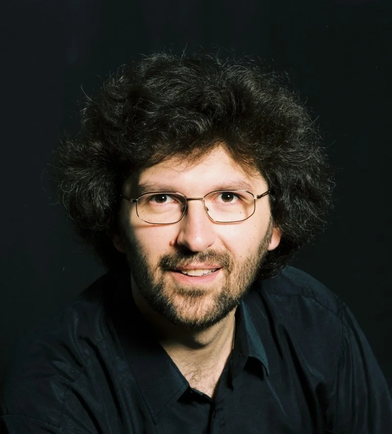 A man with curly, dark hair and glasses, wearing a black shirt, smiles at the camera against a dark background.