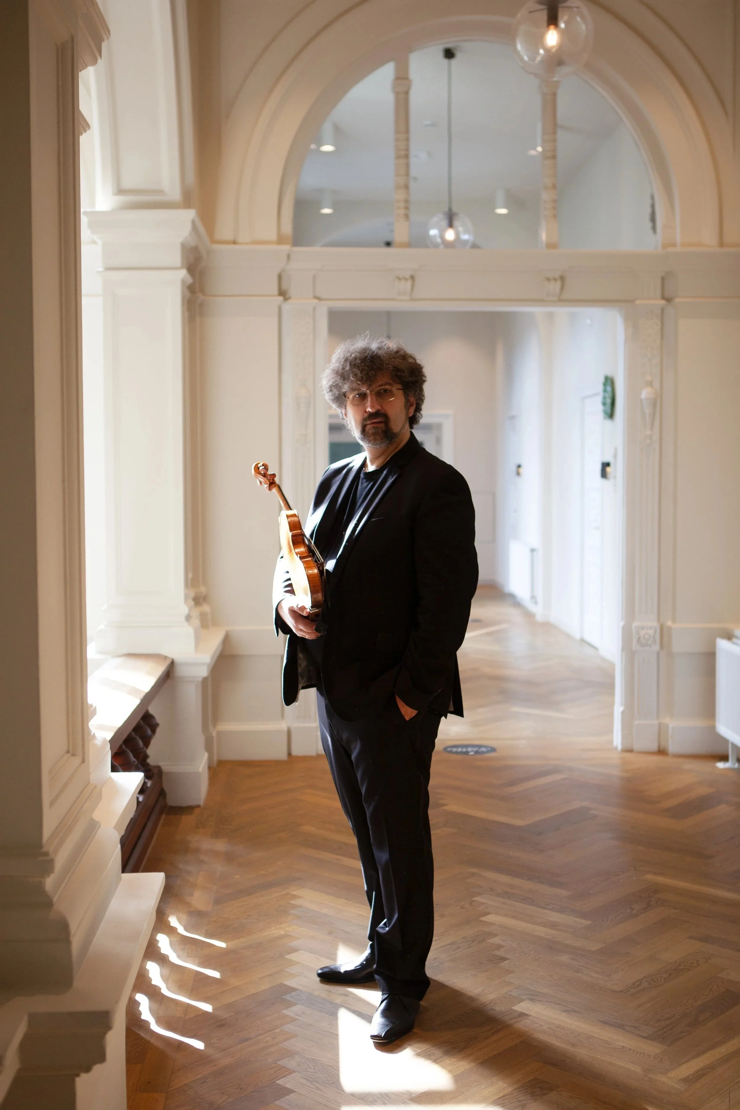A man with curly hair, glasses, and a beard, dressed in a black suit, holding a small string instrument, standing in a well-lit hallway with arched doorways, white walls, and wooden flooring.