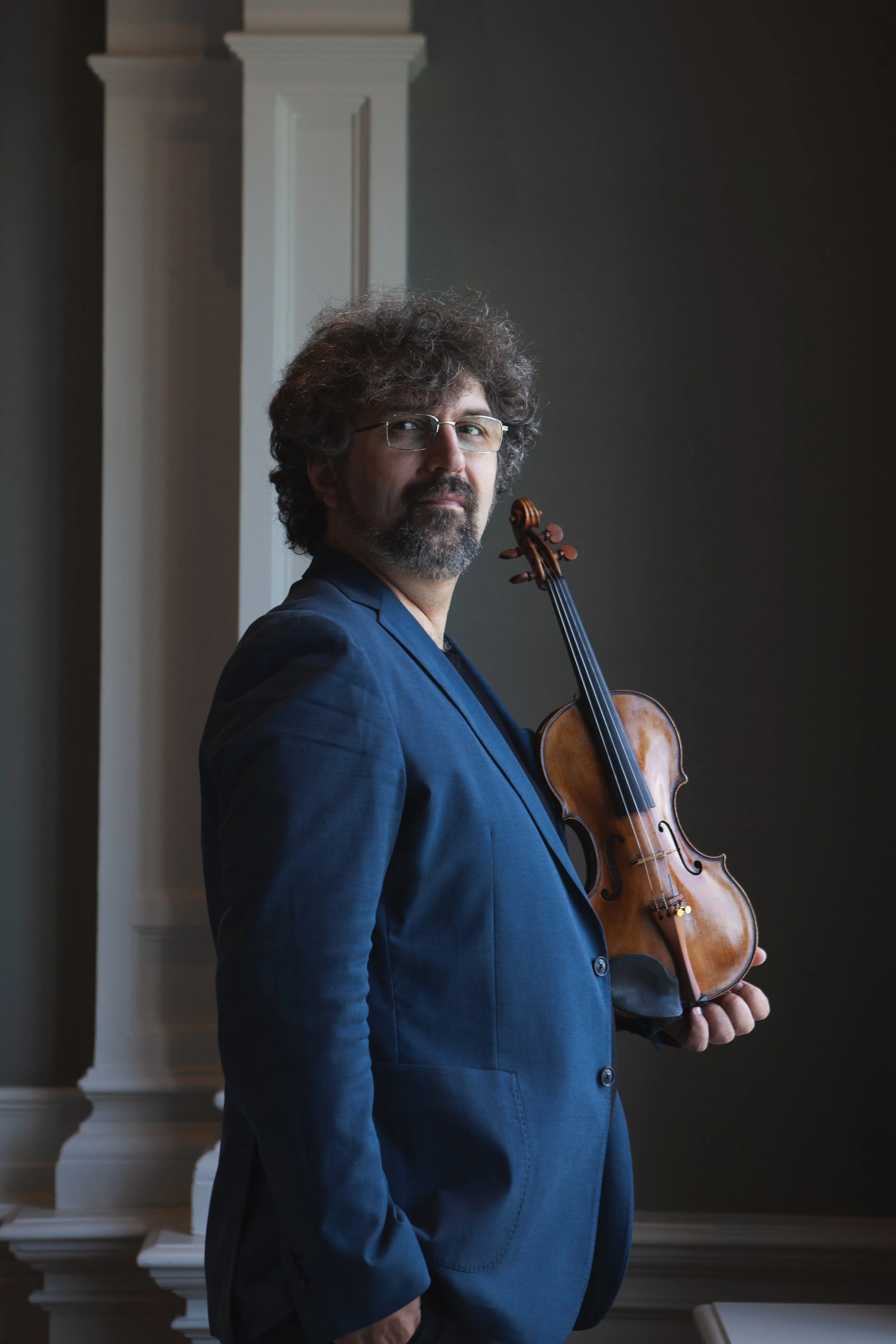 A man with curly hair, glasses, and a beard, dressed in a blue suit, holding a violin, standing indoors near a white column and a gray wall.