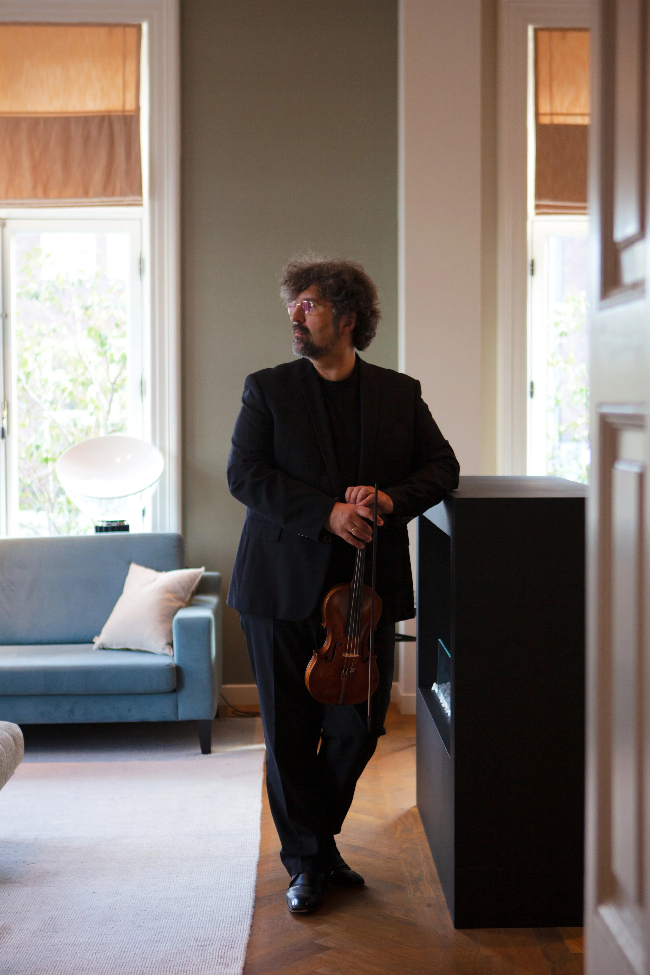 A man with curly hair and glasses, dressed in black, holding a violin, standing in a living room near windows with wooden blinds, a gray sofa with white and beige pillows, and a white chair with a side table.