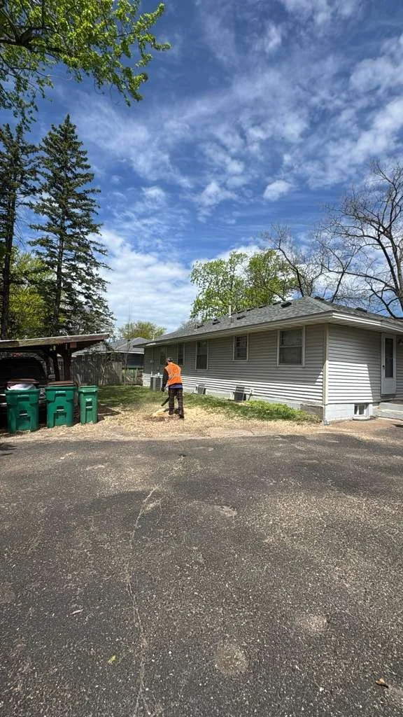 A person wearing an orange shirt and dark pants raking leaves in a backyard near a gray house with white siding. There are green trash bins near a wooden carport on the left. The sky is partly cloudy with blue sky and scattered clouds, and tall trees surround the yard.