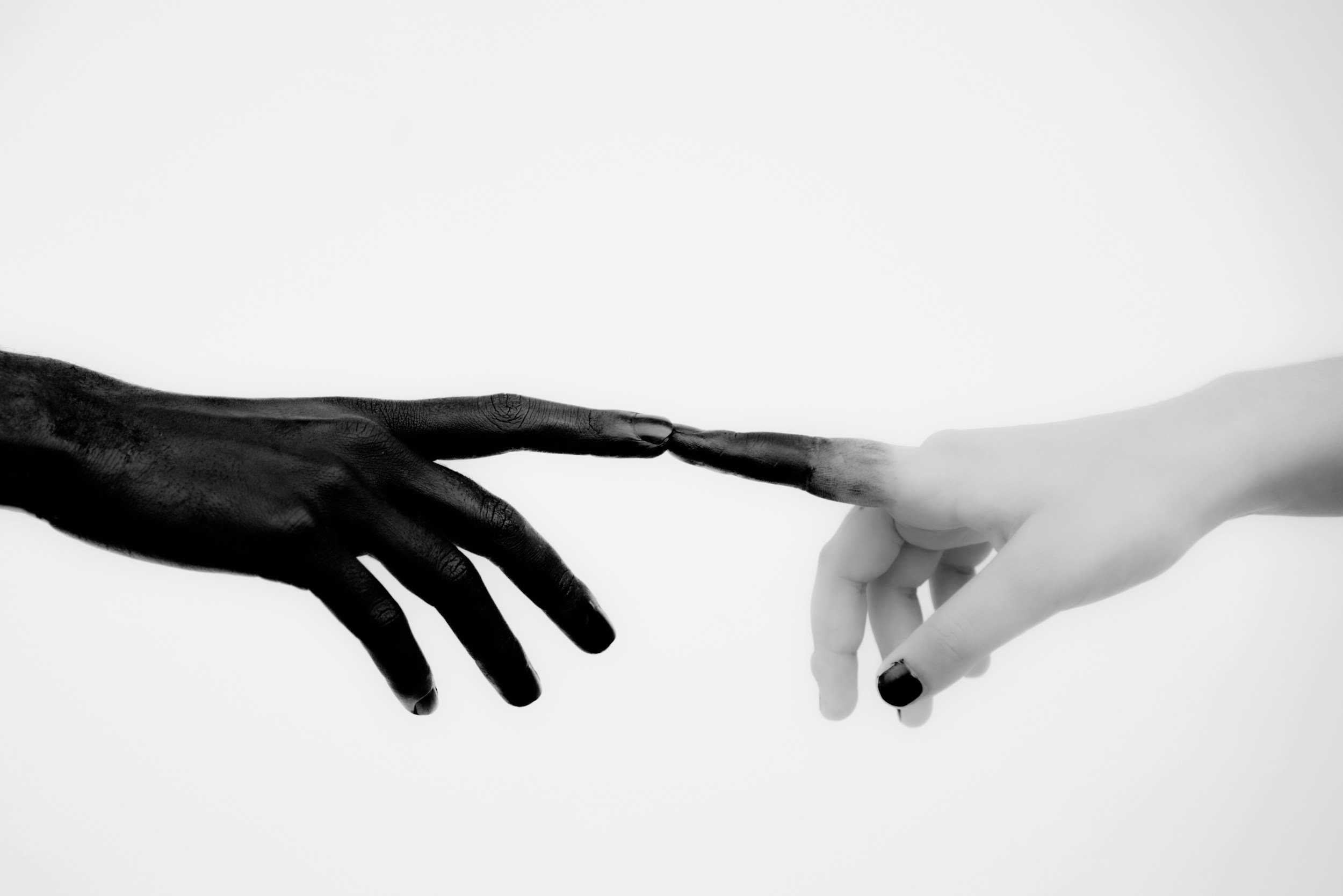 Two hands, one dark-skinned and the other light-skinned, reaching towards each other with fingertips touching against a plain background.