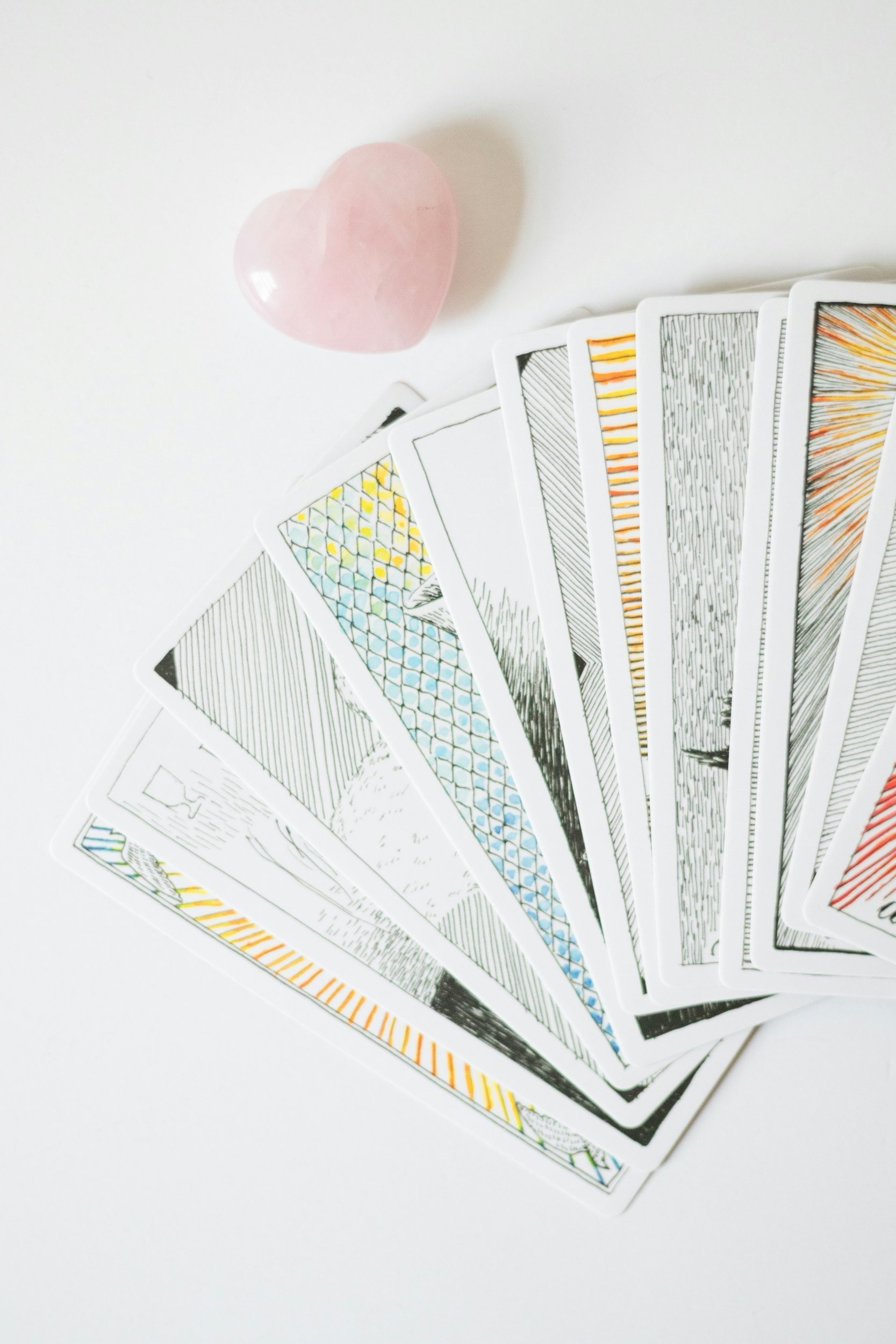 A pink, heart-shaped object next to a fan of playing cards with black and orange designs on a white surface.