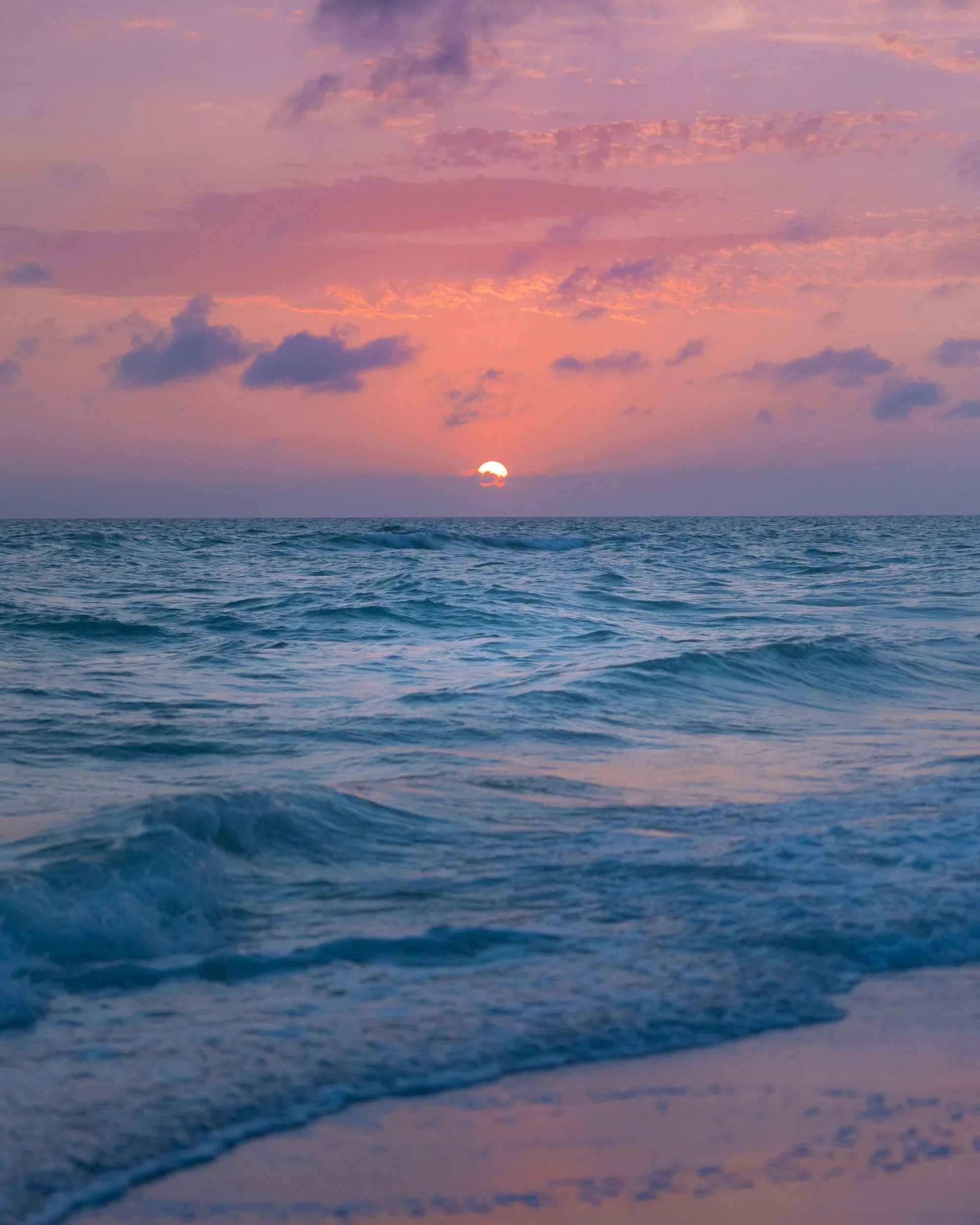 Sun setting over the ocean with a pink and purple sky and clouds.