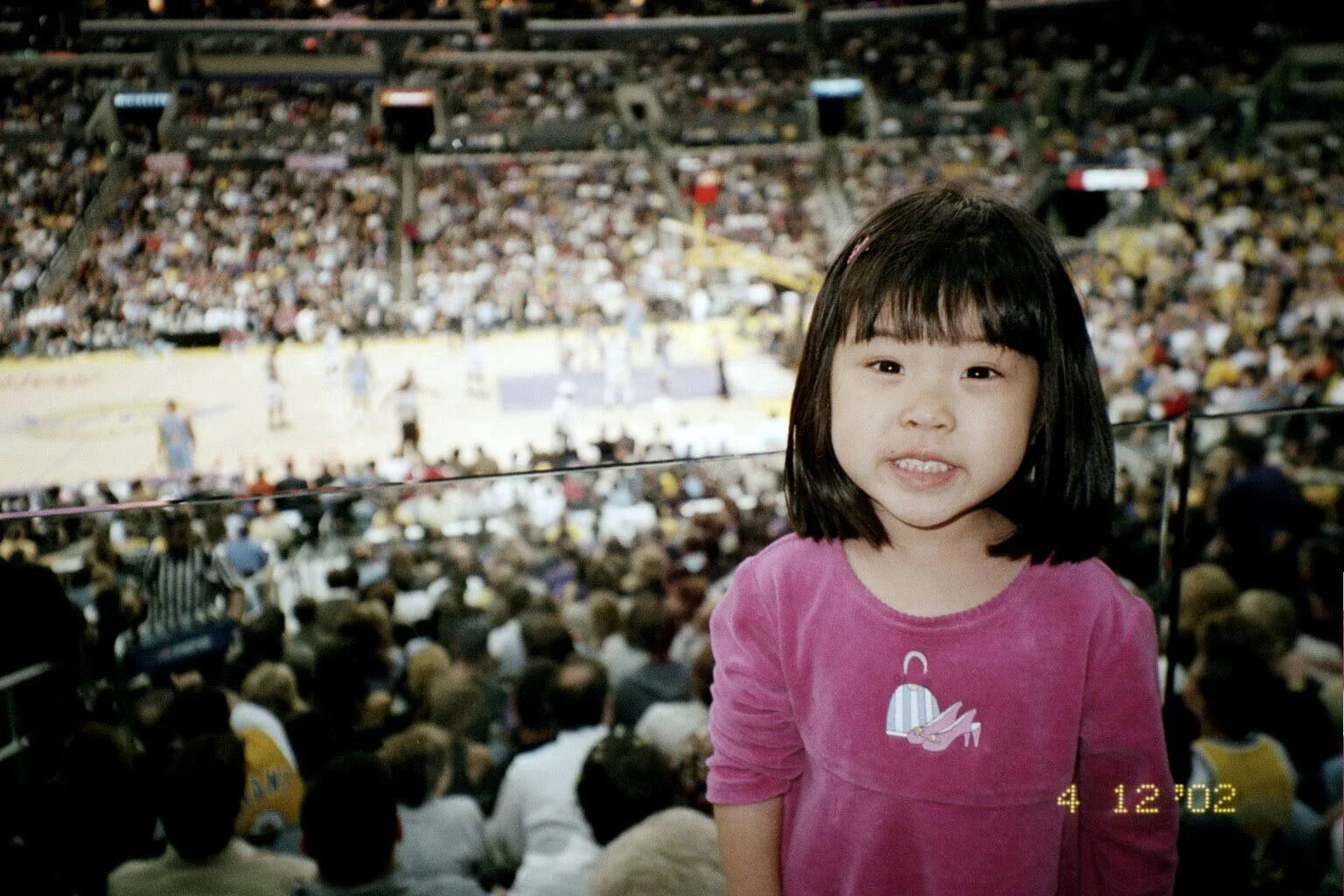 A young girl with black hair and wearing a pink shirt stands in front of a crowded basketball arena, with many spectators and players on the court in the background, and a timestamp showing 4:12:02 in the lower right corner.