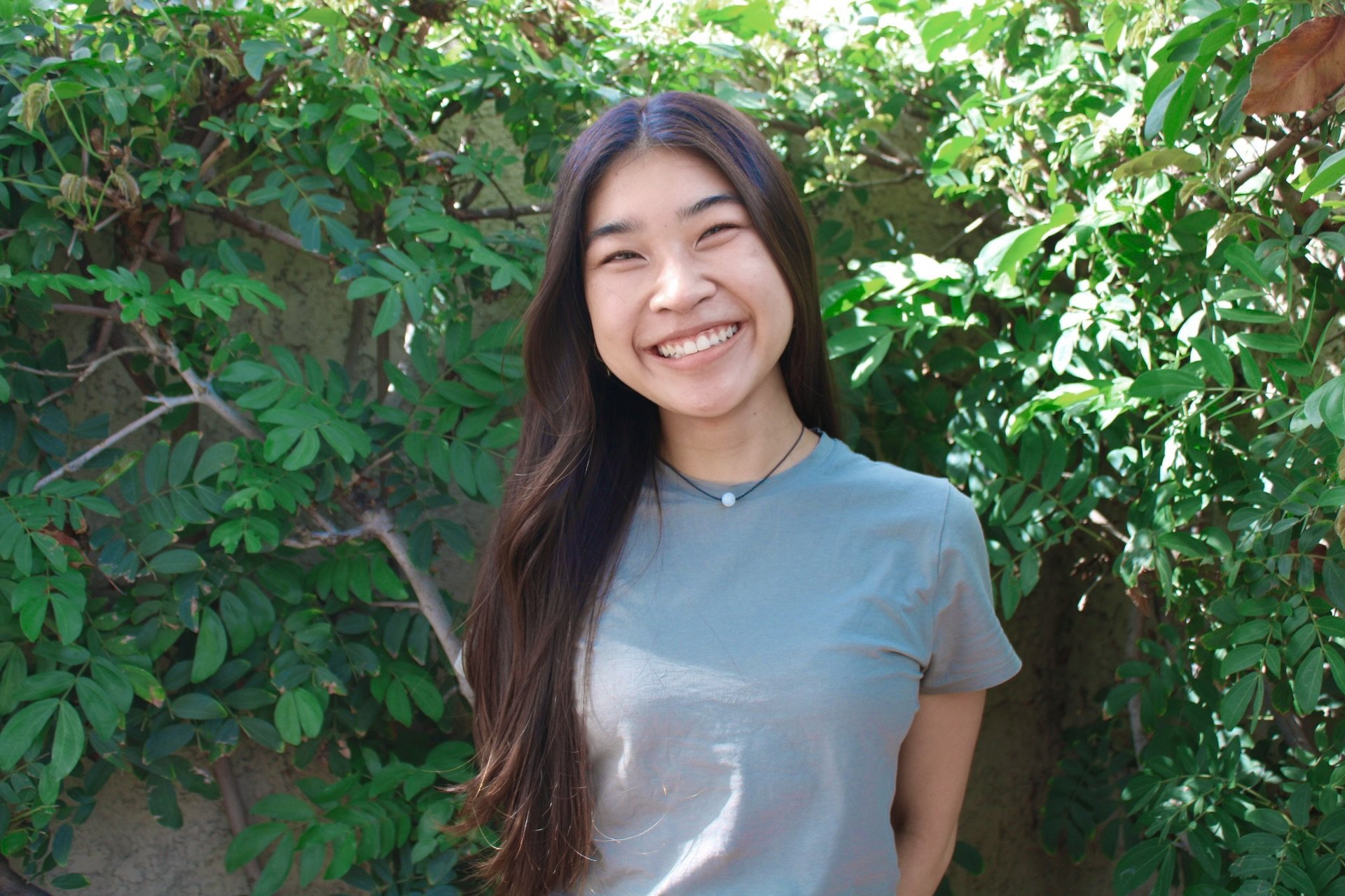 A smiling young woman with long dark hair stands outdoors in front of green leafy bushes, wearing a light gray t-shirt and a small necklace with a white bead.