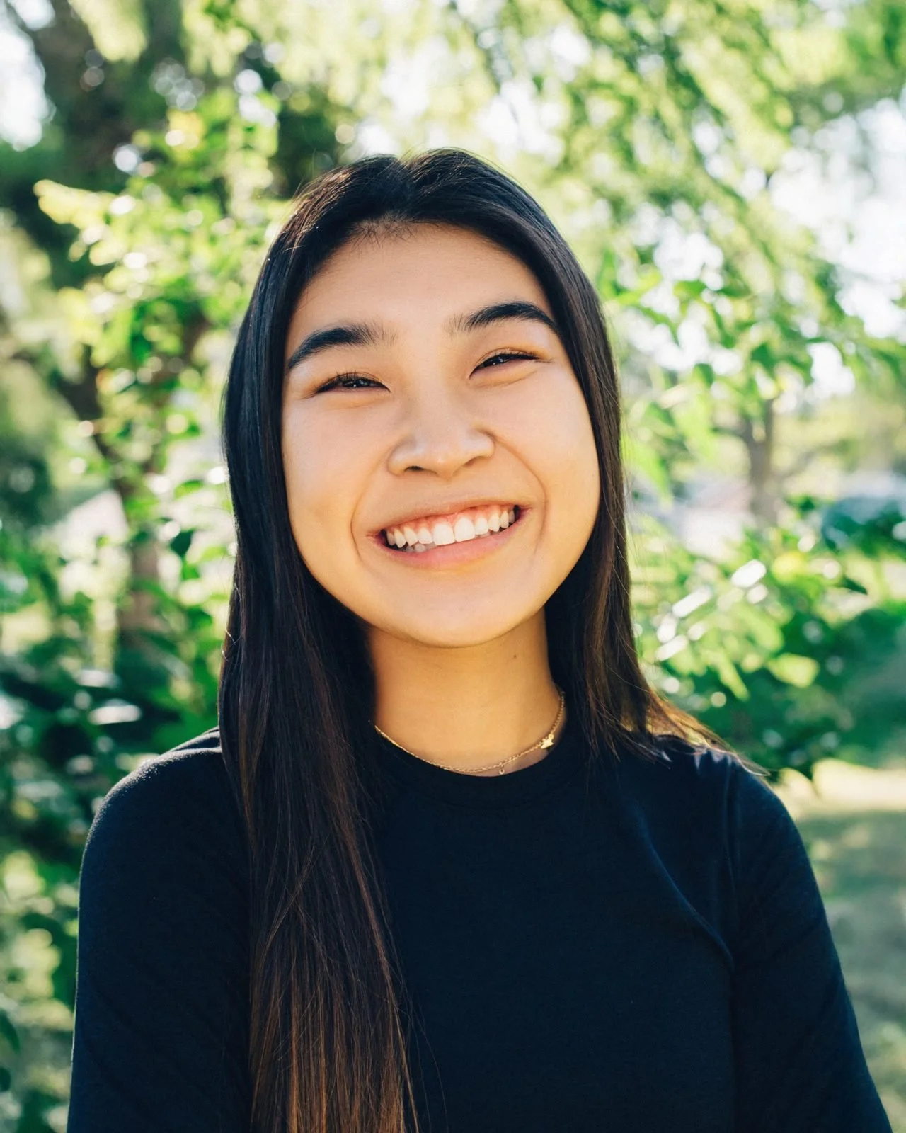 A young woman with long dark hair smiling outdoors with greenery and trees in the background.