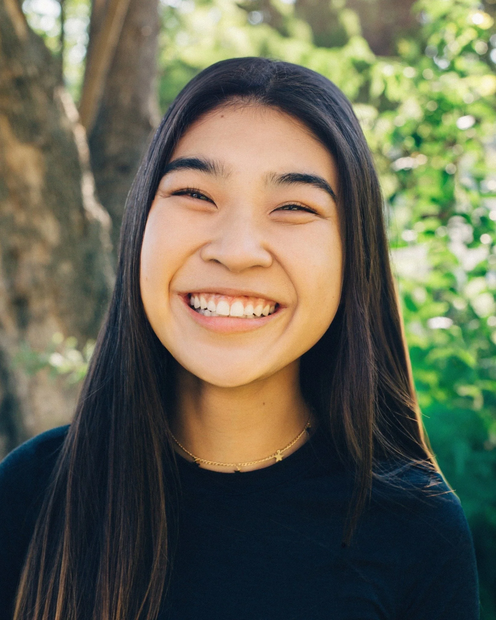Close-up of a young woman with long dark hair smiling outdoors with green trees in the background.