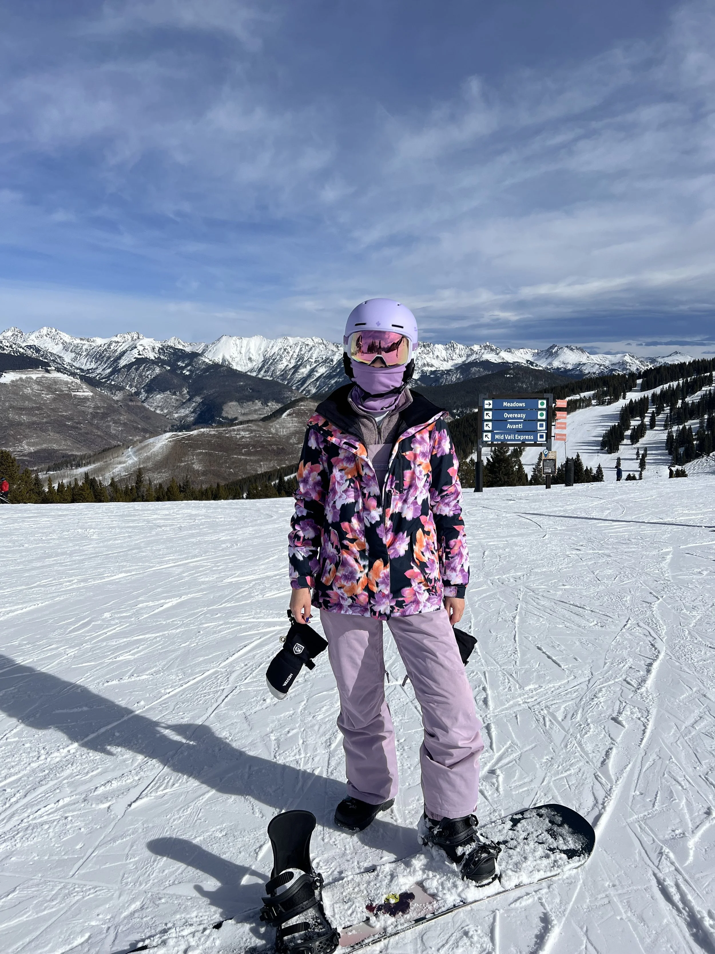 A person in a purple jacket and pink pants standing on a snowy mountain with a snowboard, wearing a white helmet and pink goggles, with snowy mountains and a blue sky in the background.