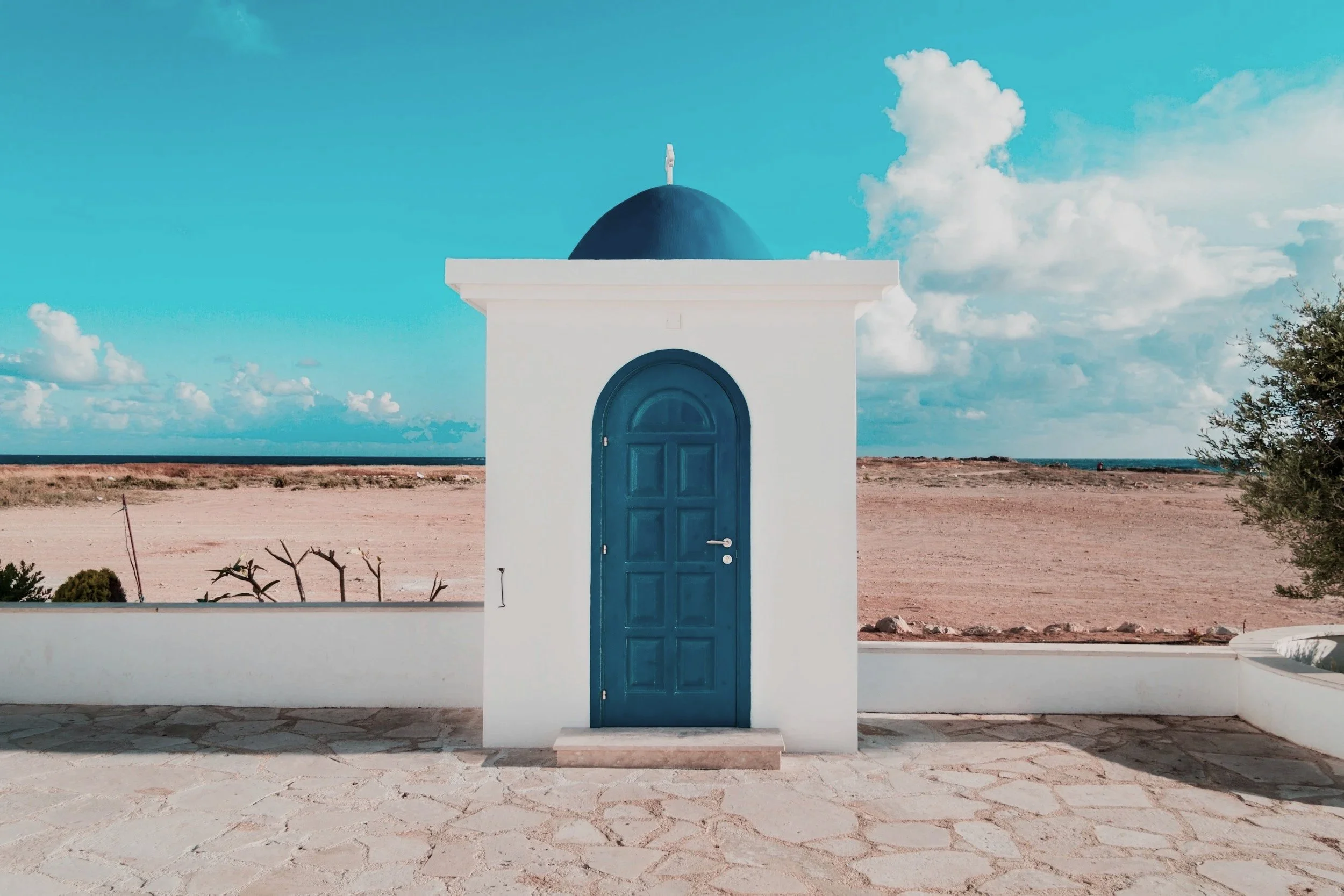 A small white building with a blue door and blue domed roof, set against a backdrop of a flat landscape with sparse vegetation and a partly cloudy sky.