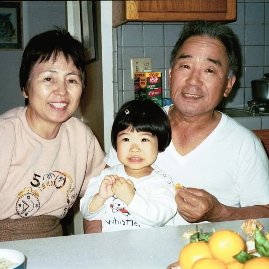 A family of three, including an elderly woman, an elderly man, and a young girl, gathered together in a kitchen, smiling at the camera. There are oranges and other items on the kitchen counter.