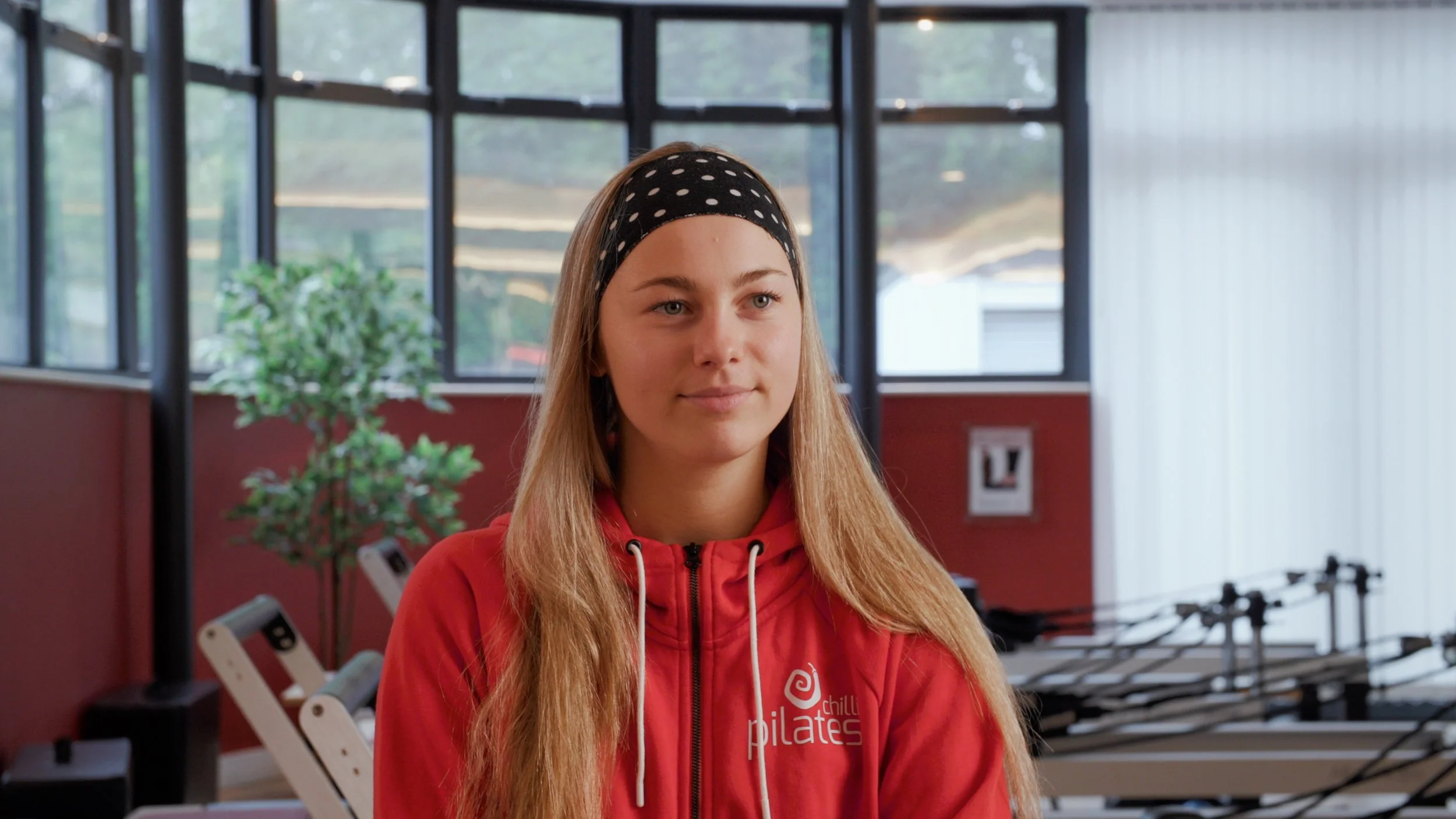 A young woman with long blonde hair wearing a black polka dot headband and a red hoodie with Pilates logo, standing inside a fitness studio with exercise equipment and large windows.
