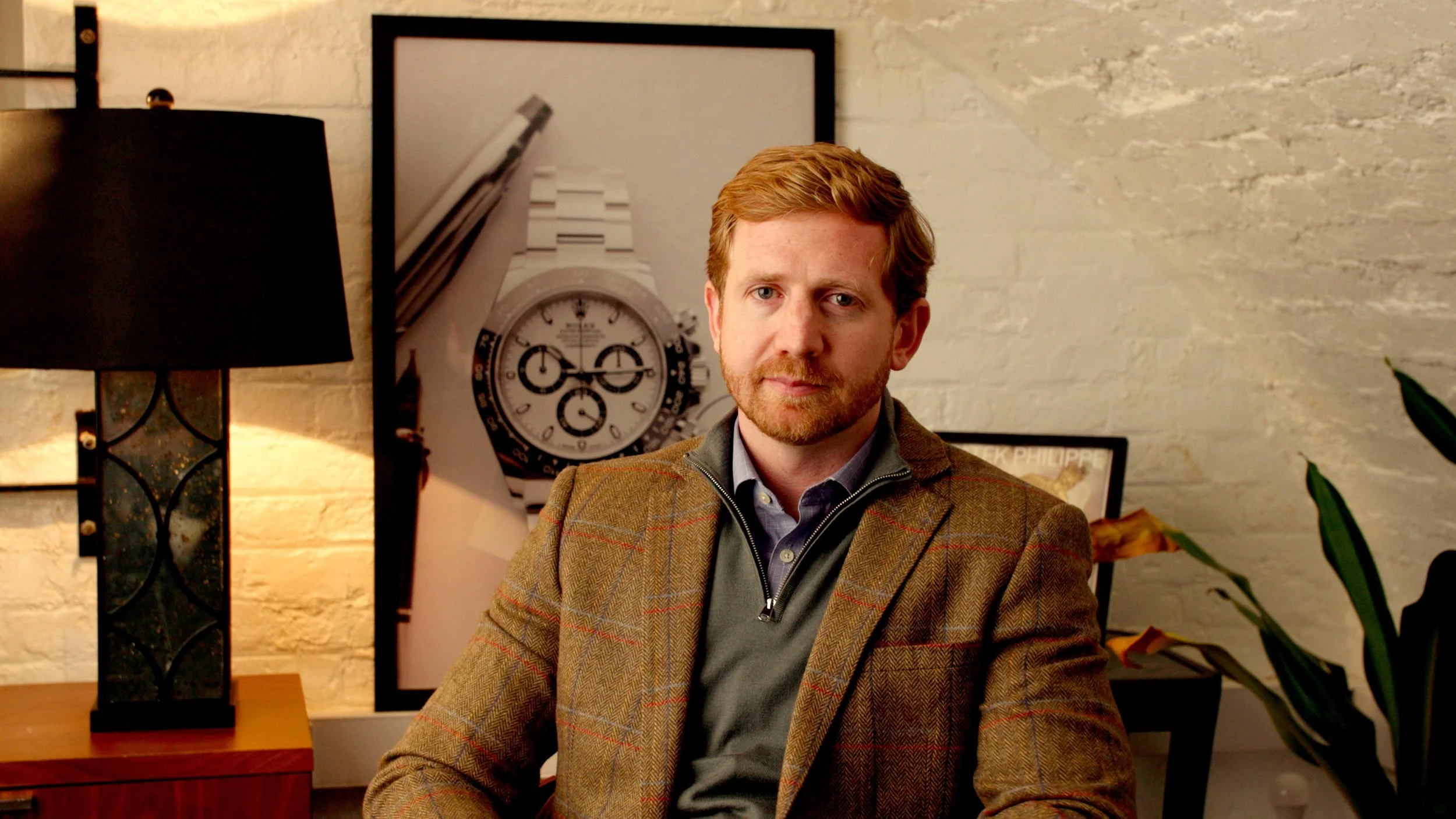 A man with red hair, beard, wearing a brown plaid blazer, seated in a room with a white brick wall, behind him a large clock picture, a black lamp on the left, and some framed artwork
