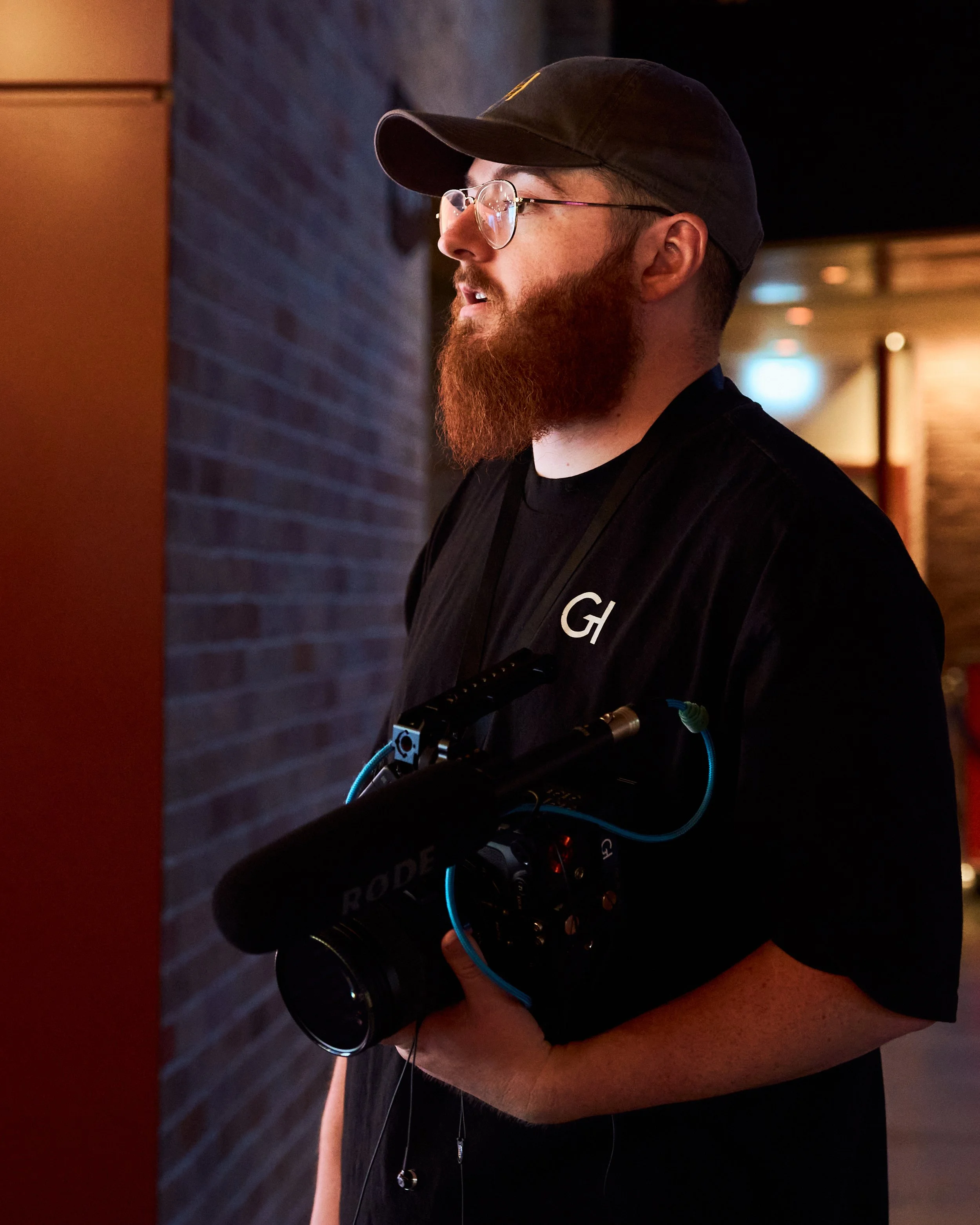 A man with a beard, glasses, and a black cap is operating camera equipment in a dimly lit setting.