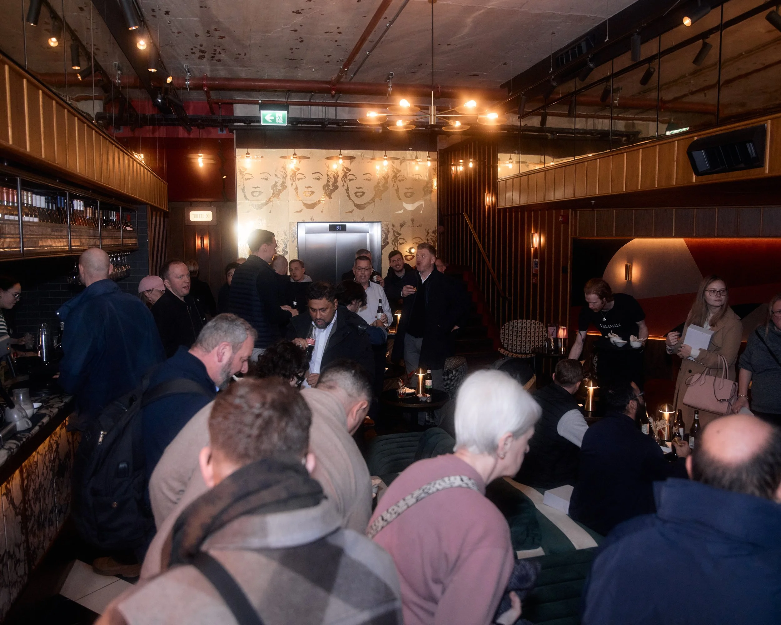 A crowded indoor event with people gathered around tables, some standing and some sitting, in a dimly lit room with warm lighting and a large mural of Marilyn Monroe on the wall behind an elevator door.