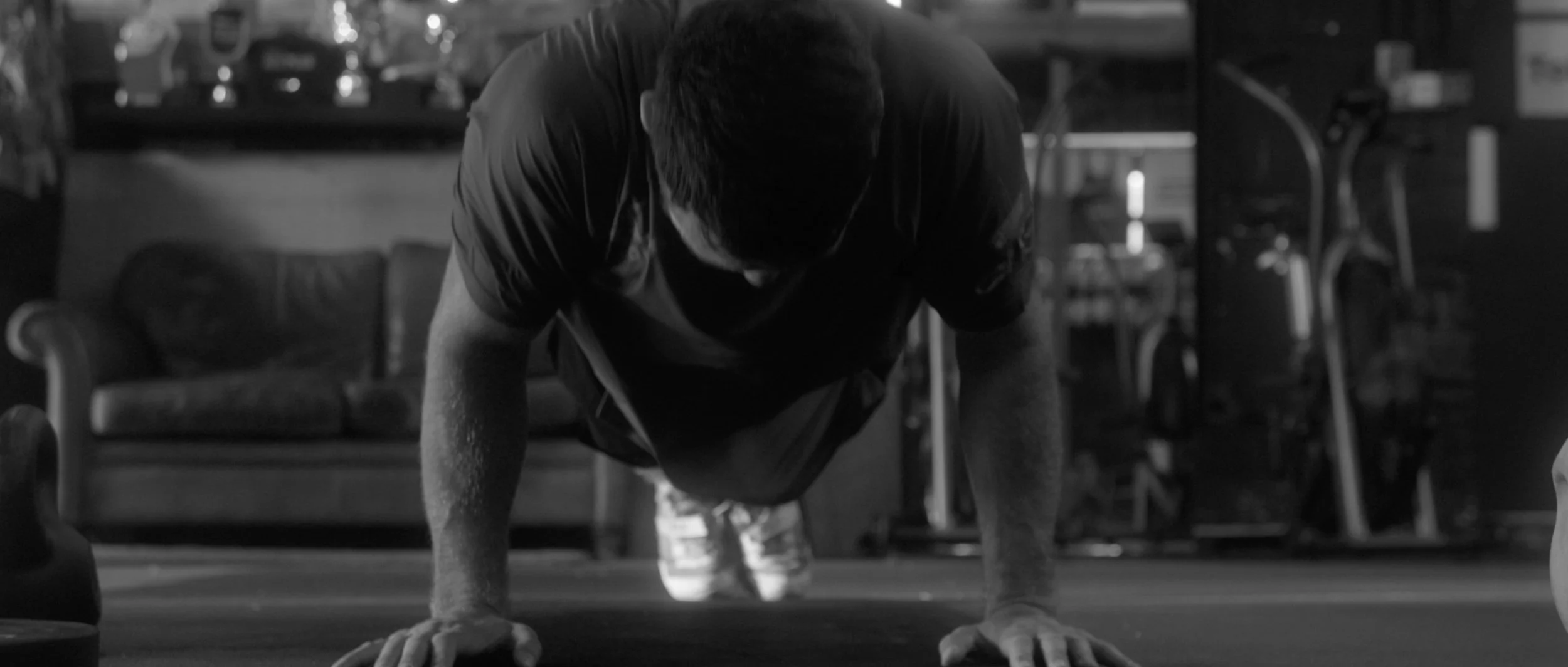 A man is doing push-ups on the floor in a gym. The background shows gym equipment and a couch.