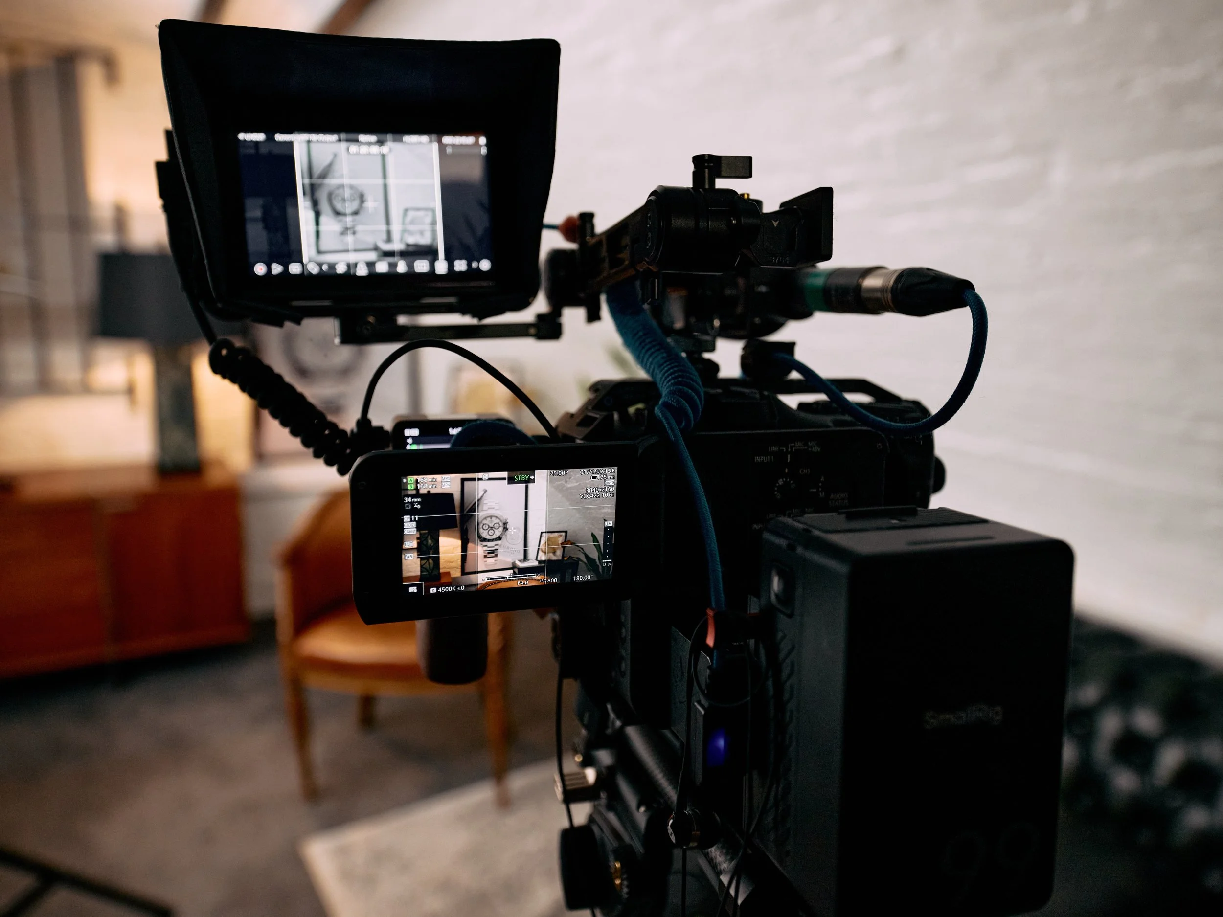The image shows a professional video camera on a tripod, recording or taking pictures inside a room with a white wall and a wooden chair in the background.