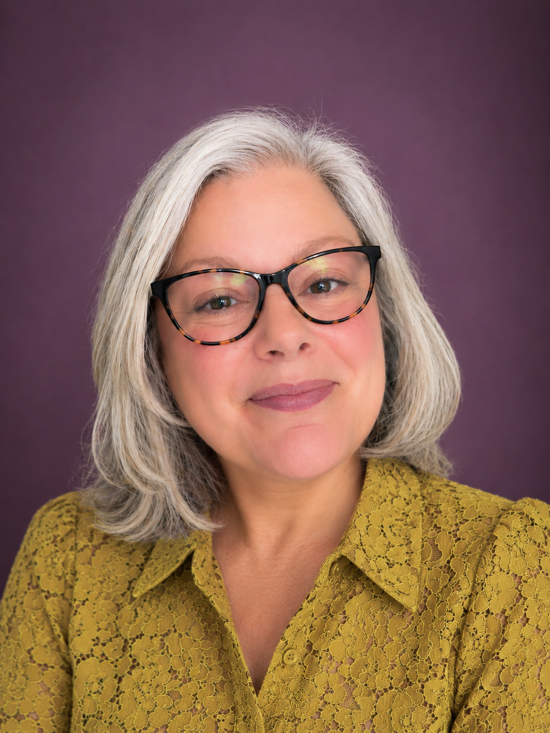 A middle-aged woman with shoulder-length gray hair and black tortoiseshell glasses, wearing a yellow lace-patterned shirt, smiling softly against a purple background.