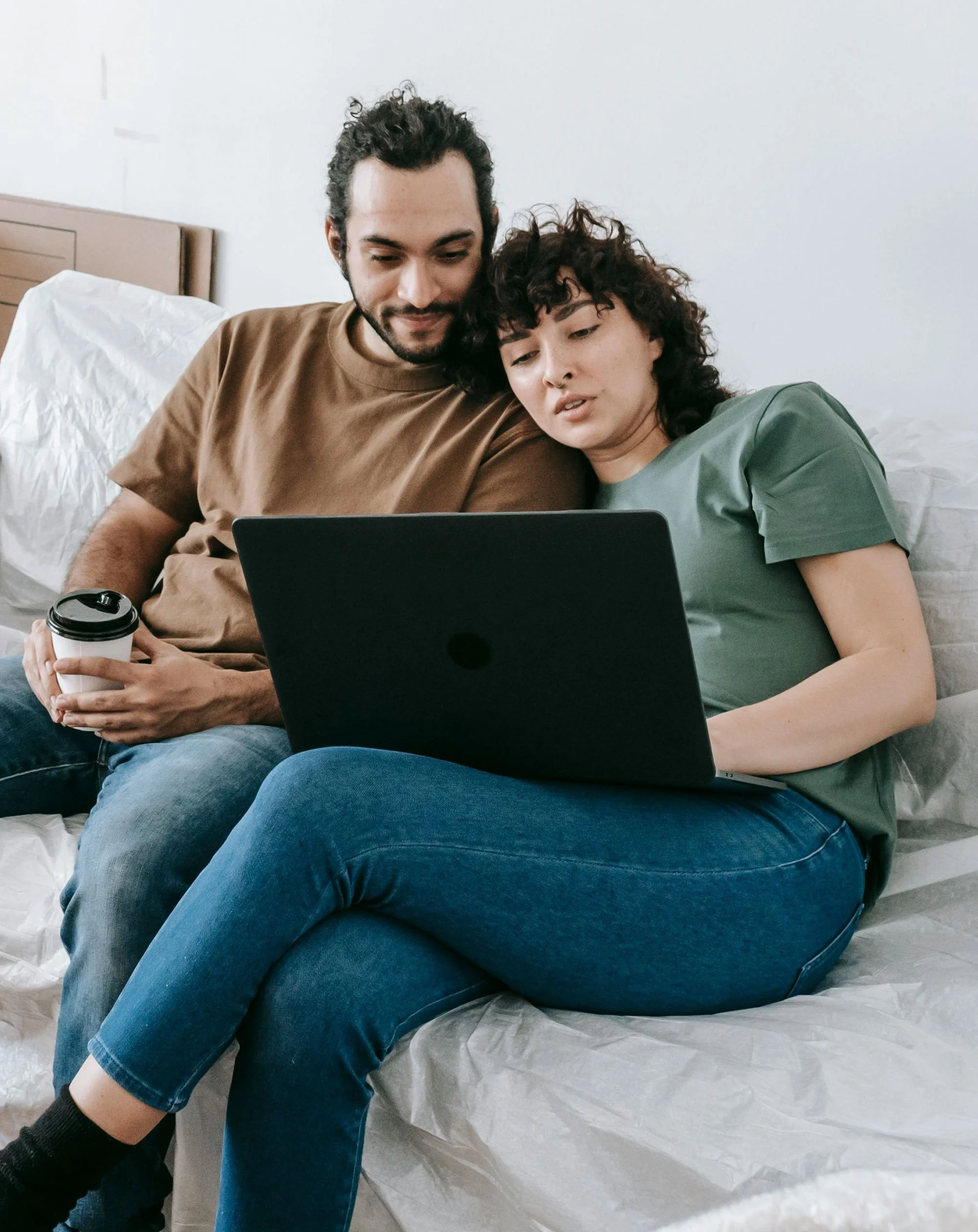 A man and a woman sit closely on a bed looking at a laptop screen together. The man holds a coffee cup in one hand. They are in a minimalistic room with a plain white wall and a wooden headboard.
