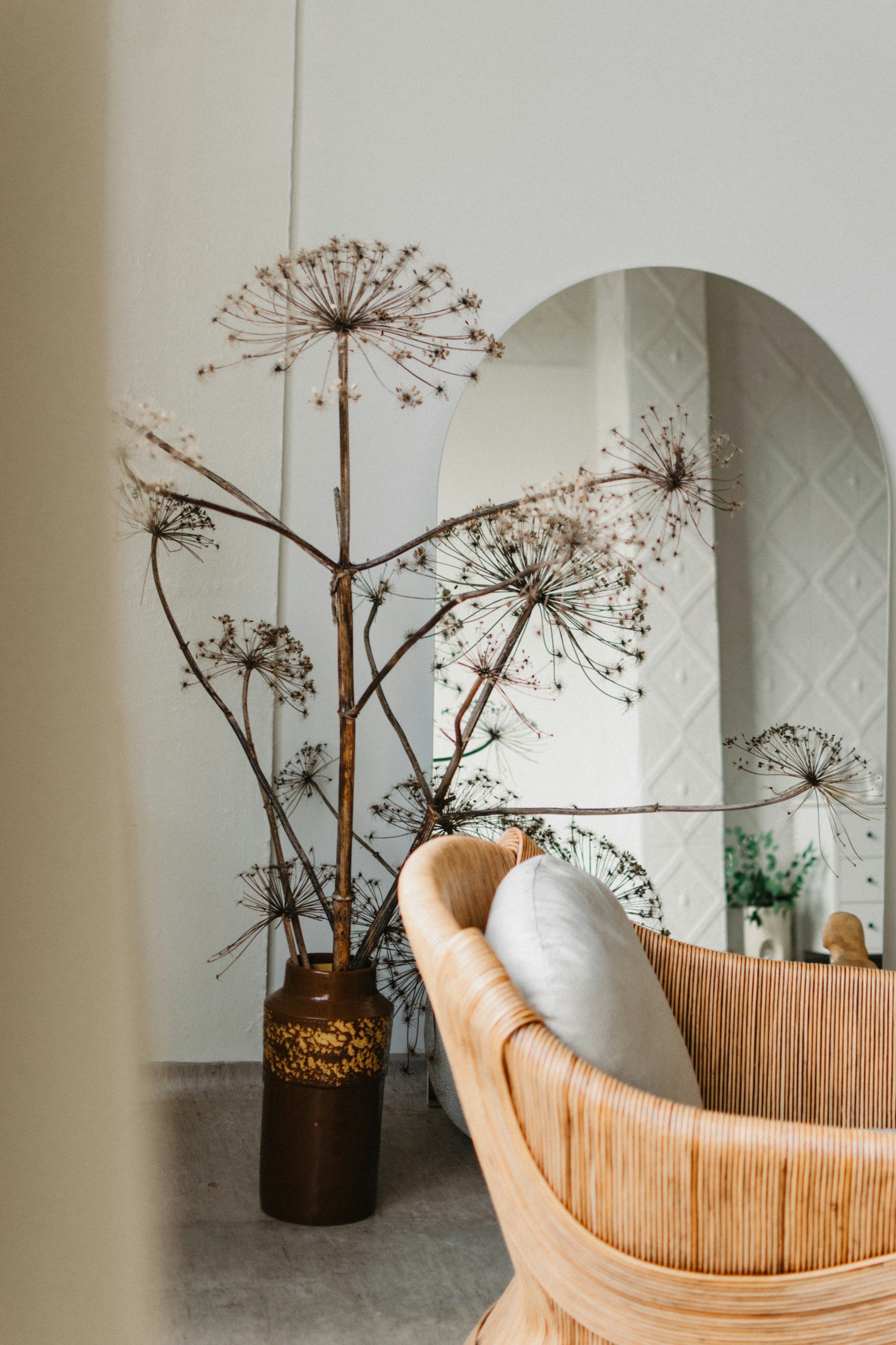 A dried floral arrangement with large seed heads in a tall dark vase, placed near a white wall with a mirror. A wooden chair with a striped fabric seat is in the foreground.