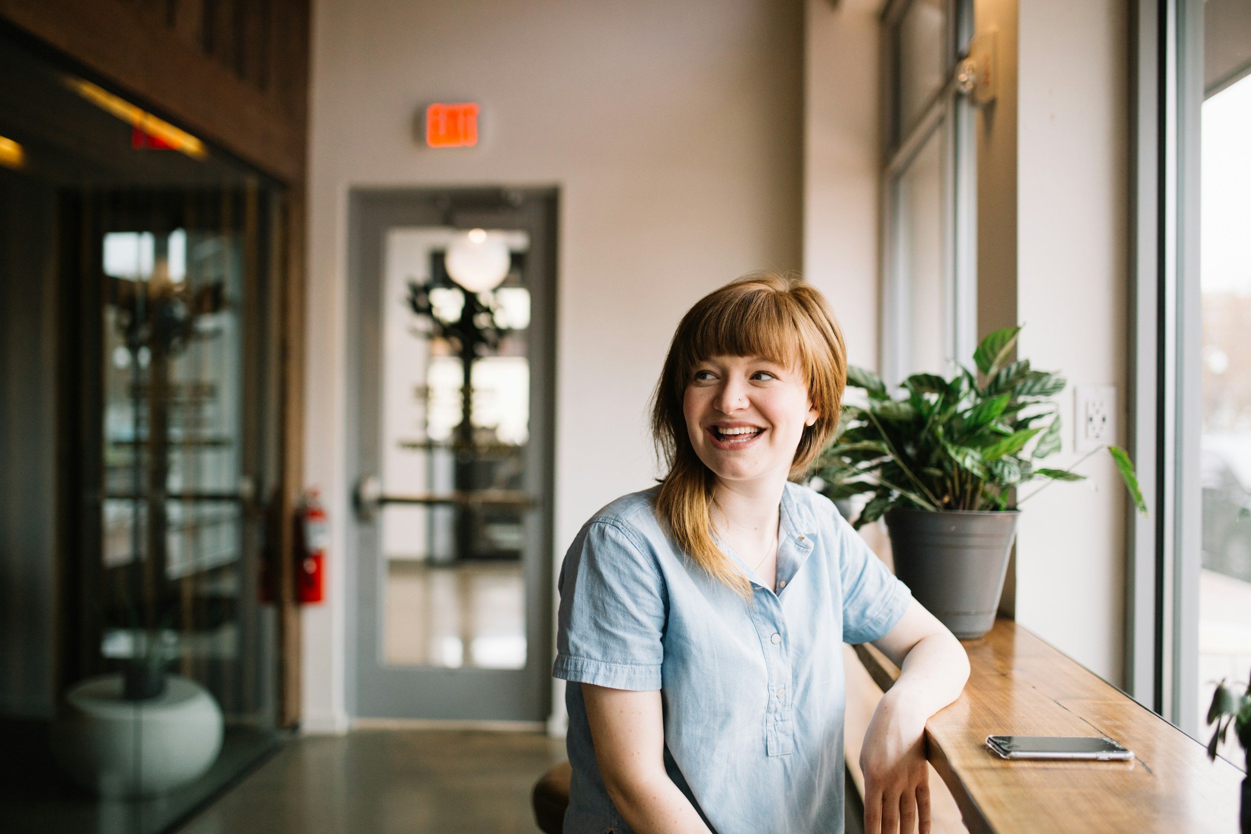 A young woman with reddish hair and bangs, smiling and looking to her left, sitting at a wooden counter near large windows with natural light, in an indoor setting with a potted plant beside her.
