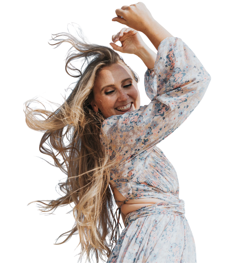 A young woman with long, flowing blonde hair is smiling and dancing with her eyes closed. She is wearing a light-colored, floral-patterned blouse and matching skirt, with her arms raised above her head in a joyful pose.
