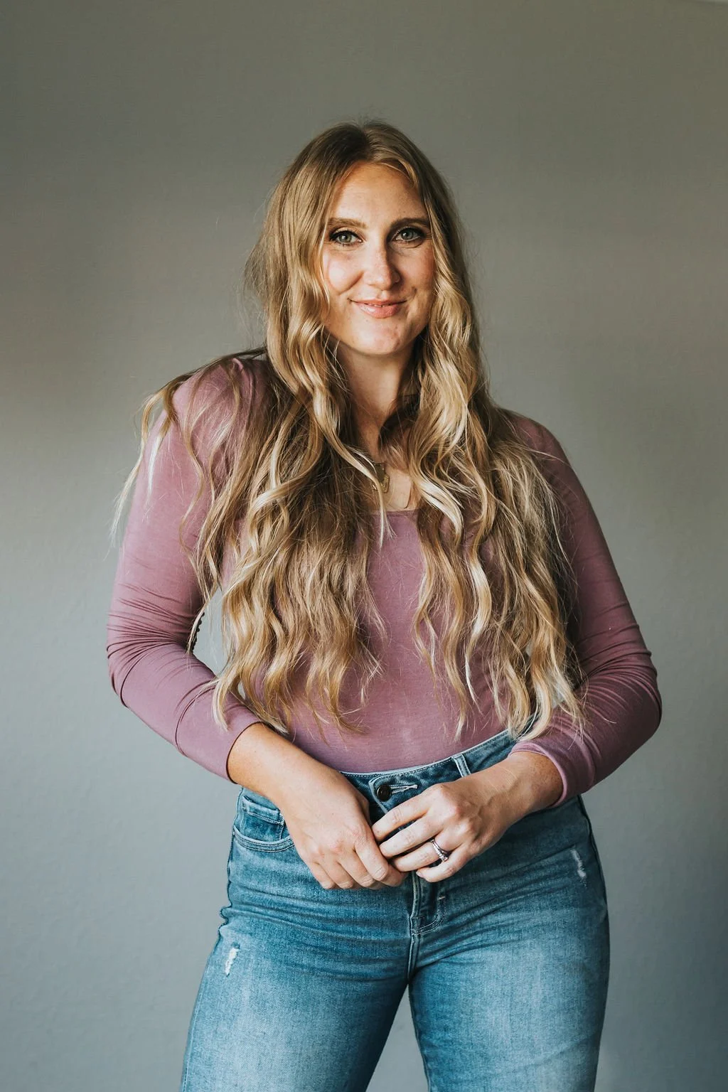A young woman with long, wavy blonde hair wearing a mauve long-sleeve top and blue jeans standing against a plain gray background, smiling at the camera.
