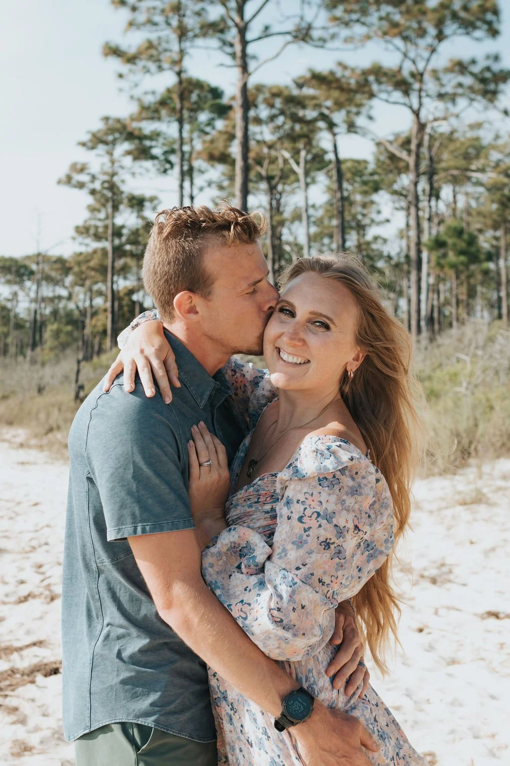 A couple on a beach, with the man kissing the woman on her cheek. The woman is smiling and wearing a floral dress, the man is wearing a short-sleeved shirt. There are trees in the background and the sky is clear.