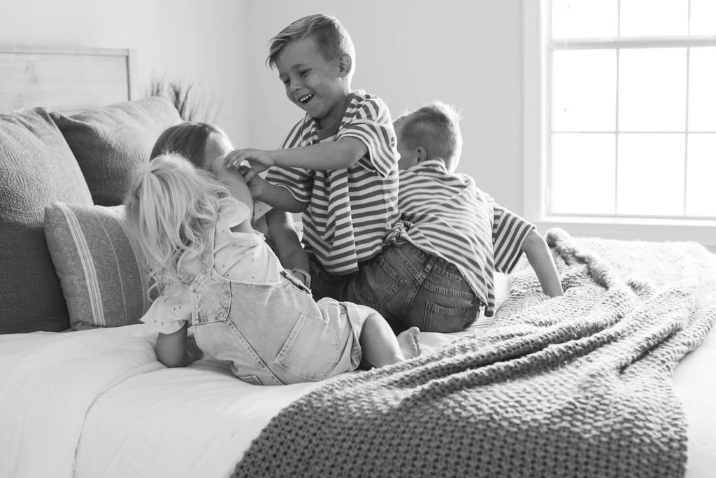Four children playing on a bed, with one girl lying down and three boys engaging with her, in a bright room with a large window.