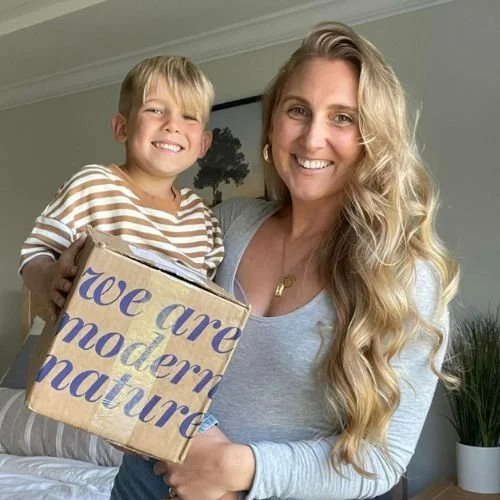 A smiling woman with long blonde hair holds a young boy with blonde hair, both holding a cardboard box that says 'we are modern nature.'