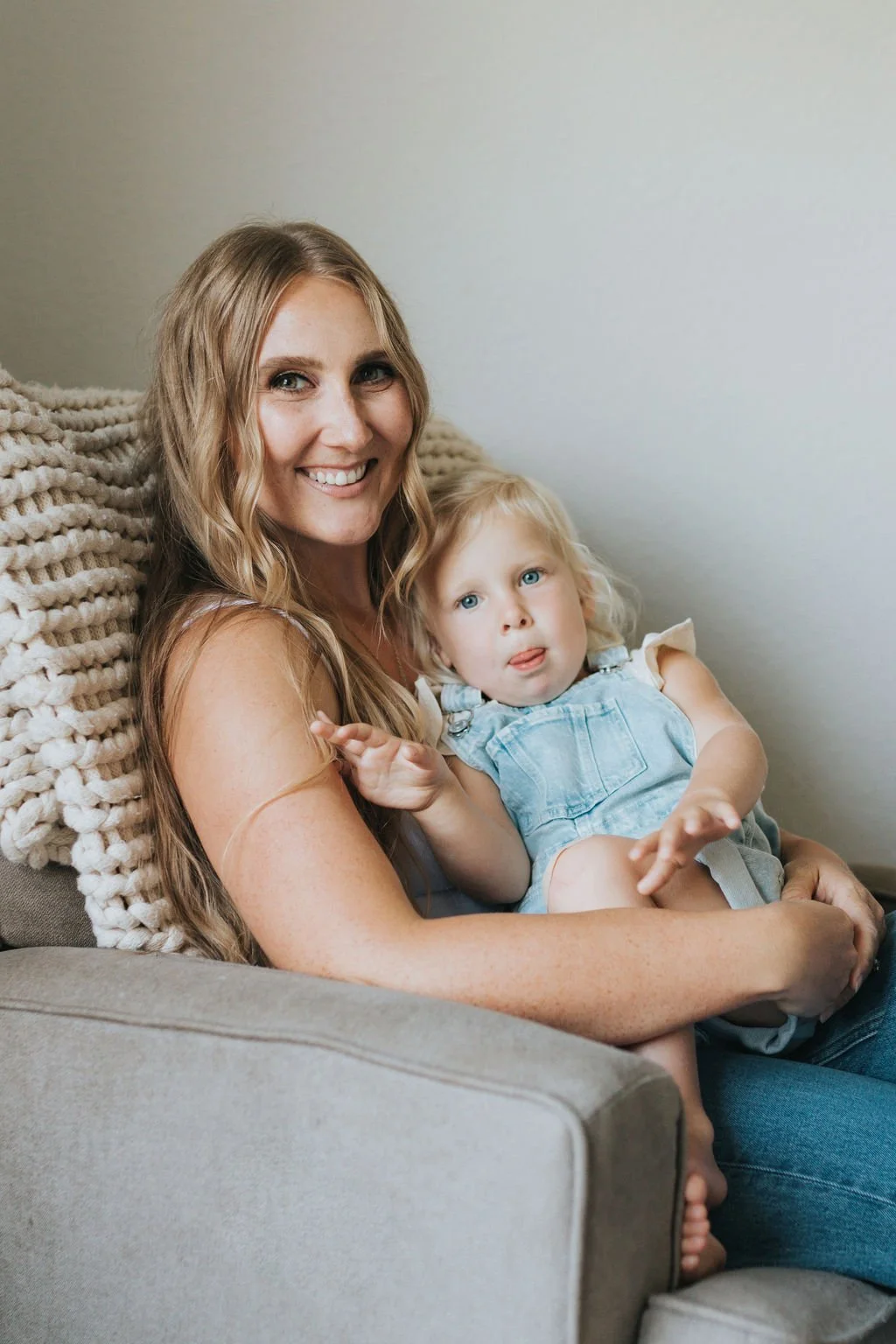 Smiling woman with long hair sitting on a beige couch holding a young girl with blonde hair and blue eyes, both looking at the camera.