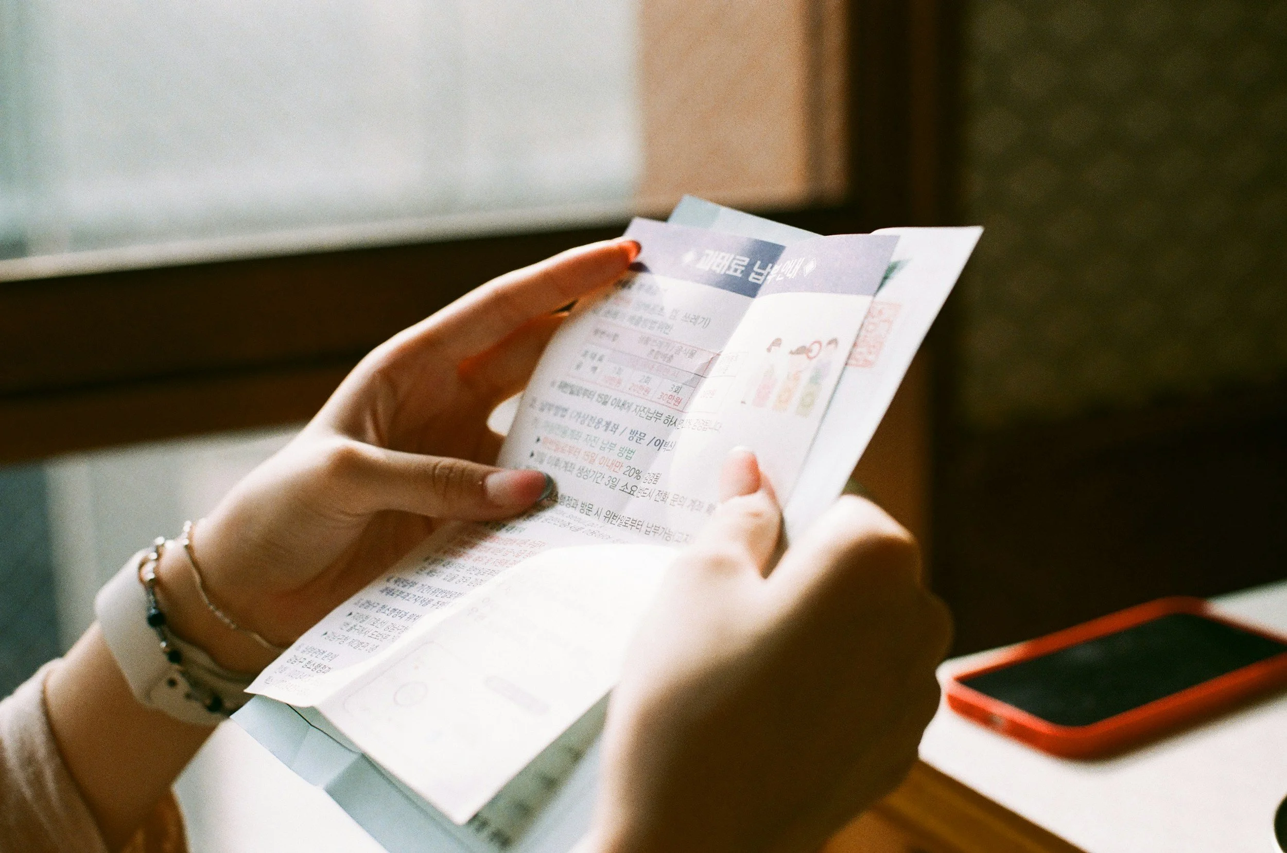 A person holding a paper with printed text and colored sections. The person is sitting at a desk, and a red device, possibly a smartphone, is also on the desk.