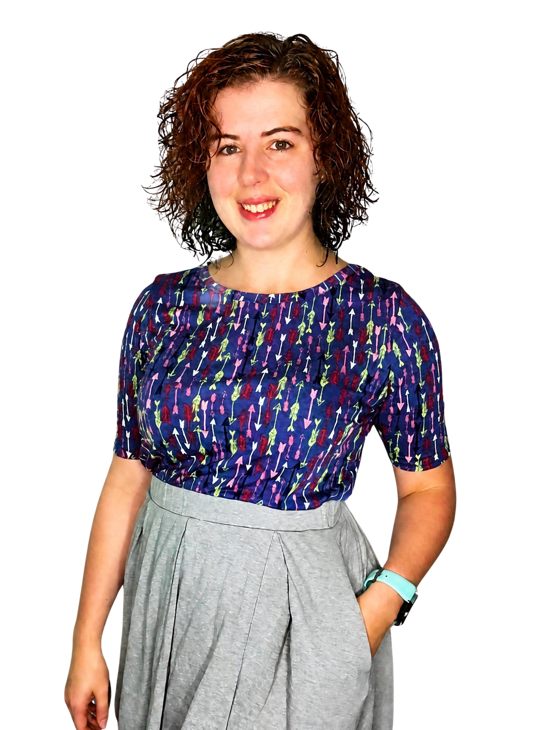 A woman with shoulder-length curly hair, wearing a navy blue short-sleeved top with colorful arrow patterns, a gray pleated skirt, and a turquoise wristwatch.