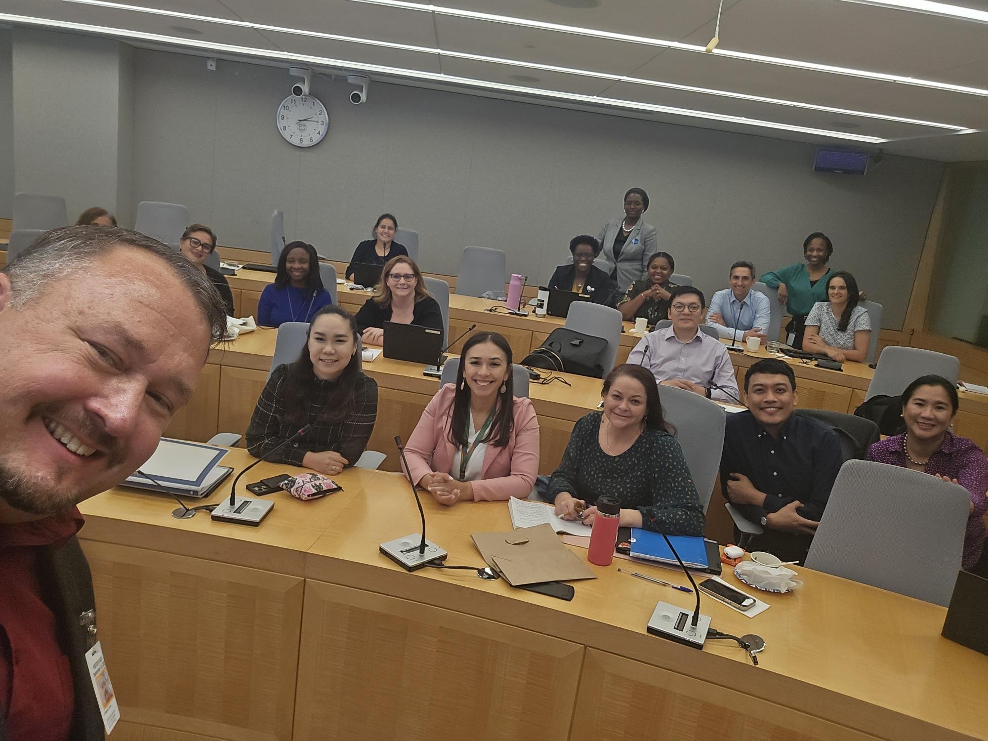 A group of diverse people in a conference room, smiling and posing for a photo. There are conference tables, laptops, notebooks, and cups on the tables. The room has a wall clock showing approximately 2:12 and a TV mounted on the wall.