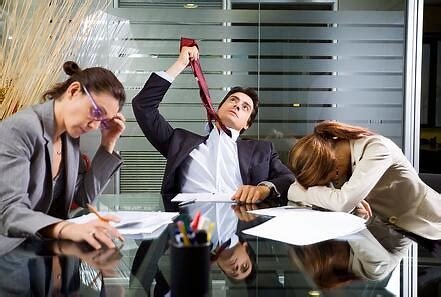 Three business professionals in a meeting room, looking frustrated; the man in the middle is stretching a rubber band, the woman on the left has her head in her hands, and the woman on the right is resting her head on the table.