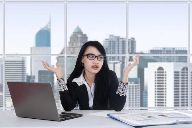 A woman with glasses and long dark hair sitting at a desk in an office, gesturing with her hands, with a laptop and open book on the desk, large windows showing a city skyline in the background.