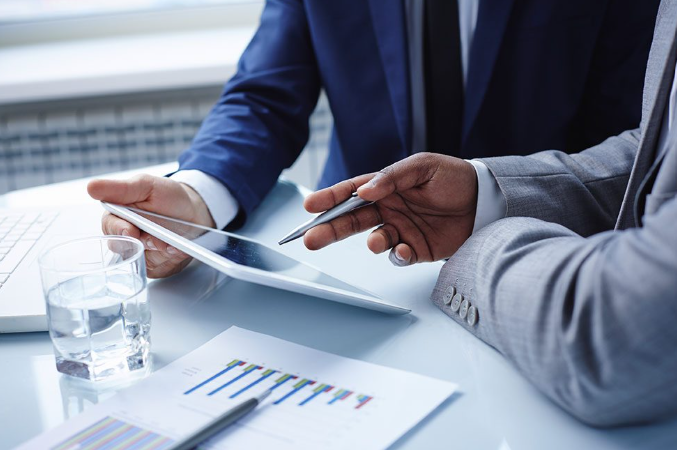 Two people in business suits working together at a desk with a tablet, a glass of water, and financial charts.