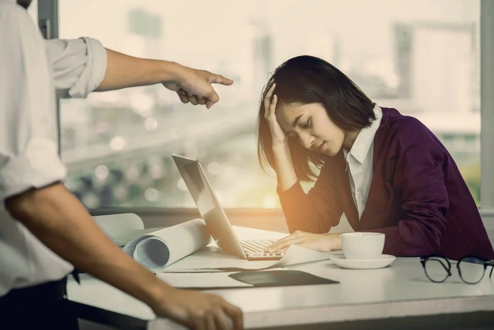 A woman sitting at a desk holding her head in frustration while looking at a laptop. A man is pointing at her, standing nearby. The scene appears to be in an office setting.