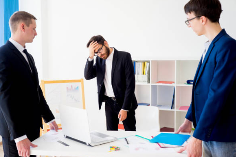 Three men in business suits are in a meeting room with a white wall and bookshelf in the background. One man appears stressed or frustrated while the other two look on.